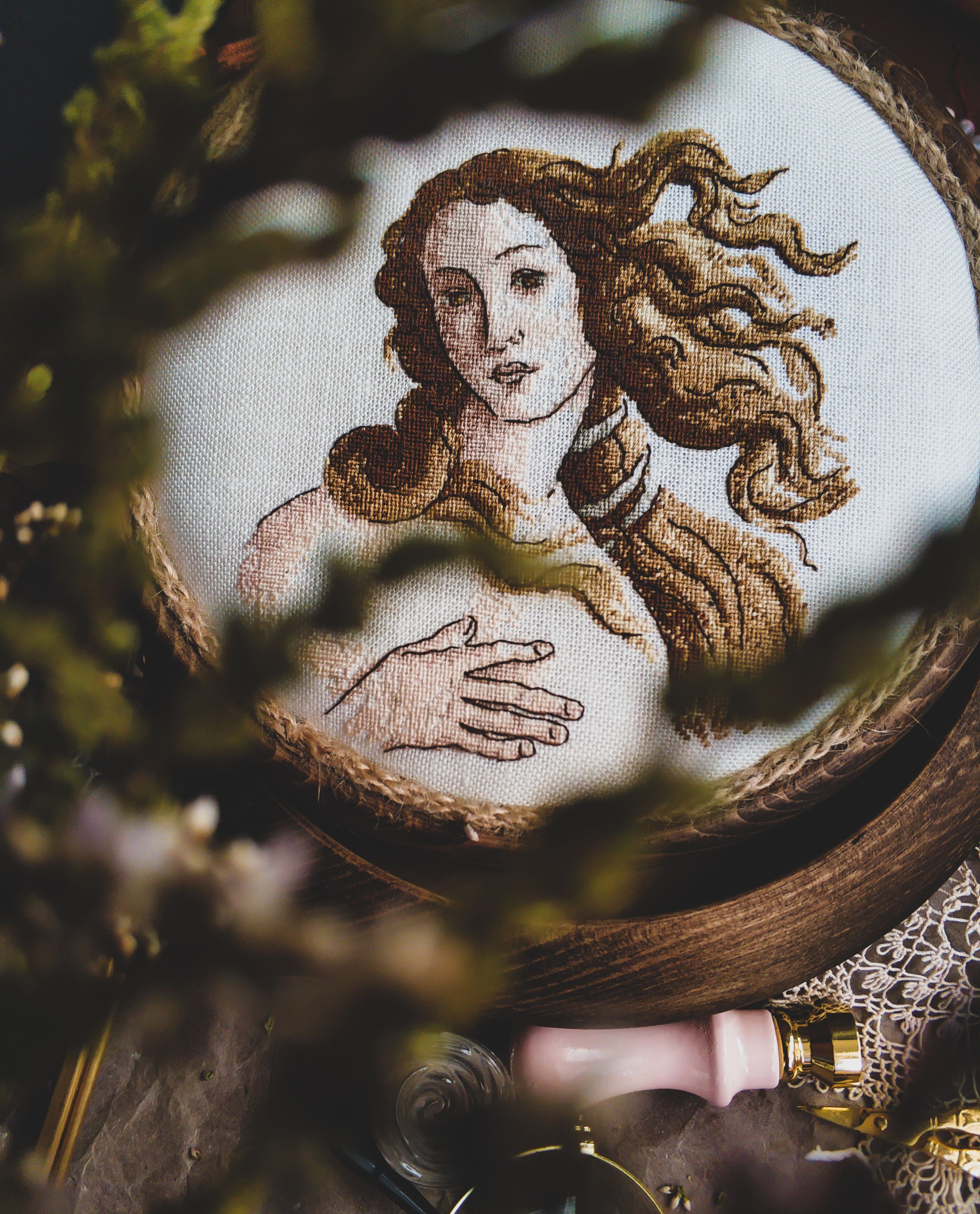 Close-up of an embroidered portrait of a woman with long, flowing hair, on a white fabric with a wooden frame around it. The embroidery is detailed, showing her face and part of her arm, with some greenery and decorative objects partially visible in the foreground and background.