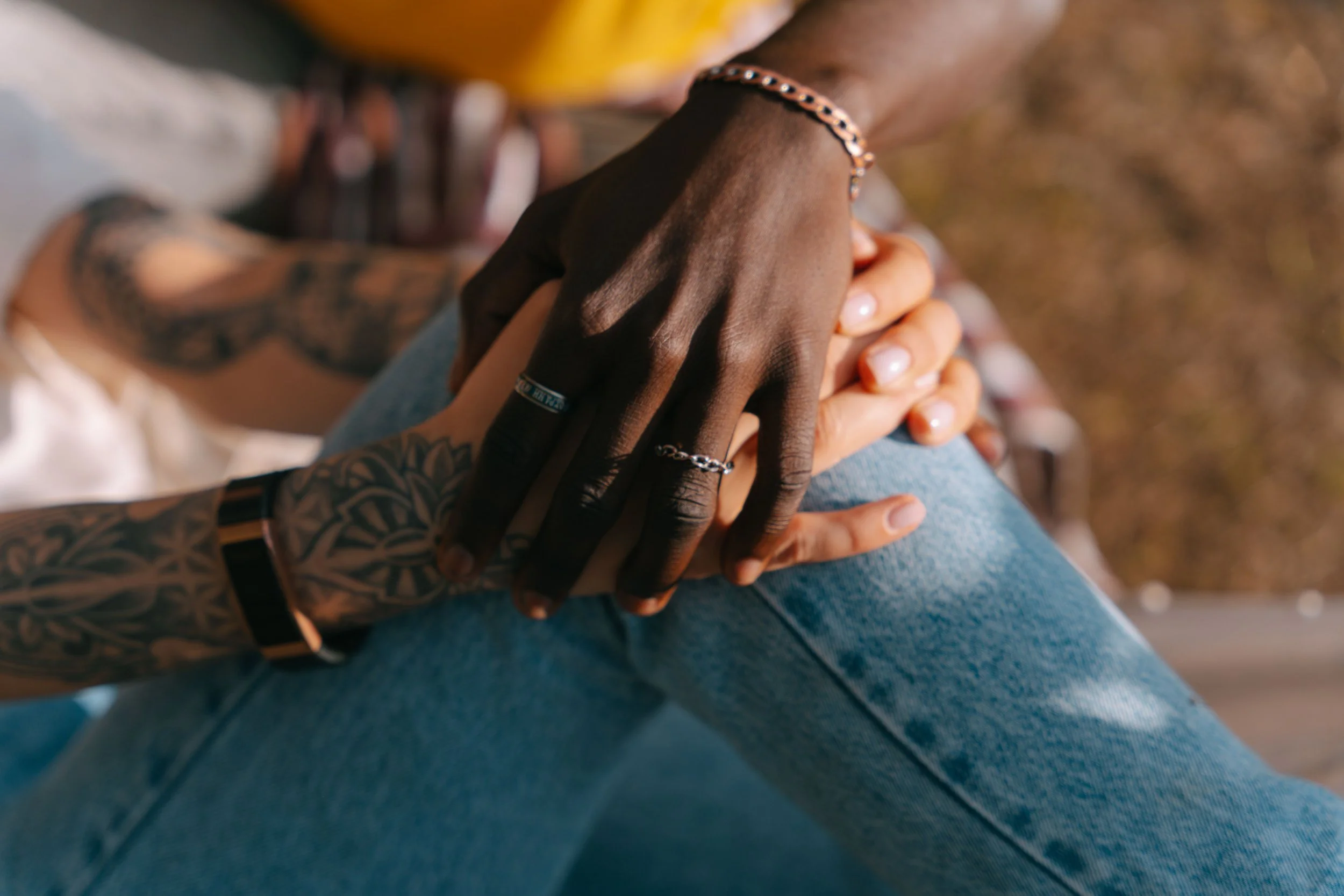 Close-up of two people holding hands, with one person's hand resting on top of the other's. One person has darker skin and is wearing a bracelet and ring, while the other has lighter skin. They are sitting outdoors.