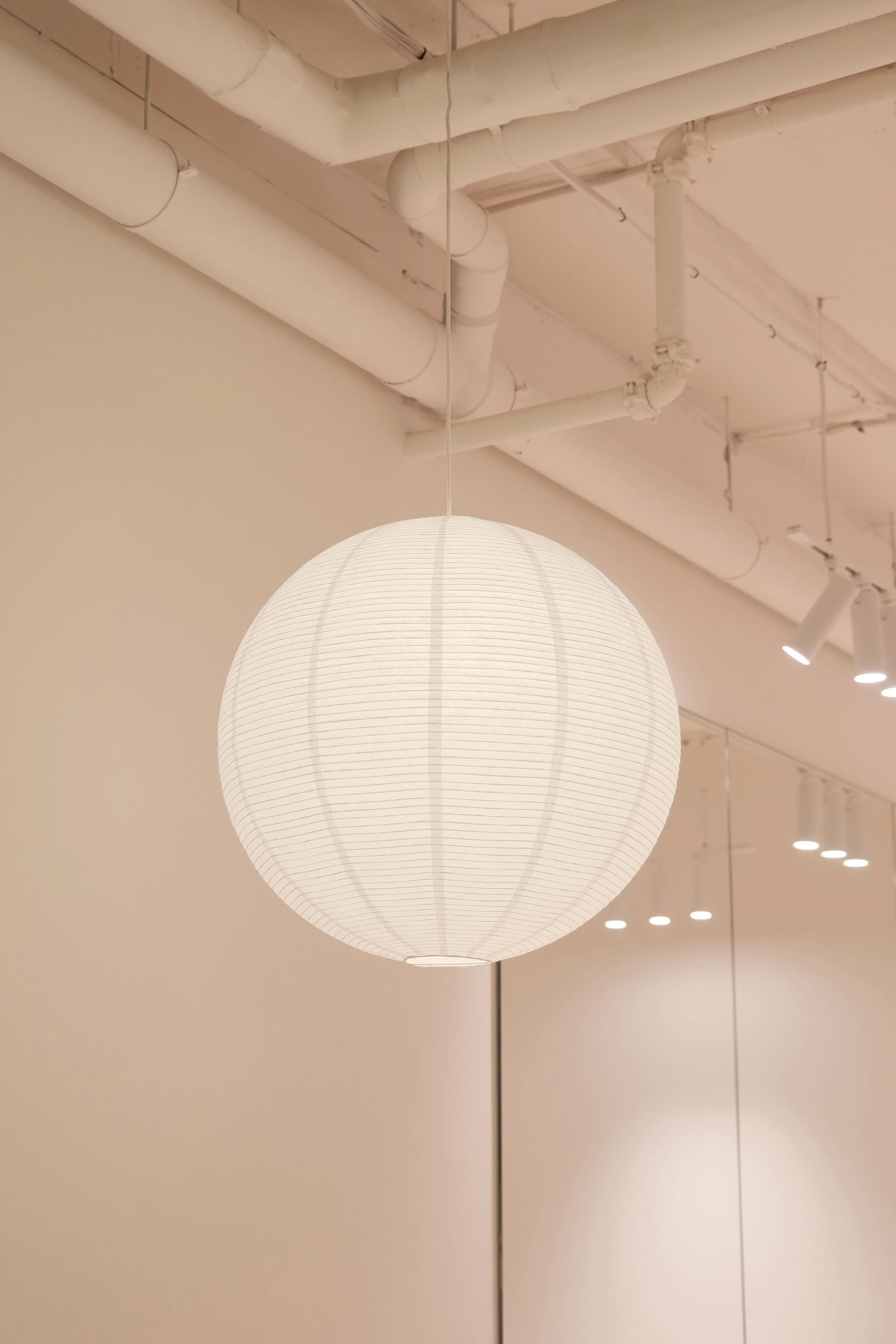 A white paper lantern hanging from a ceiling with exposed pipes and modern track lighting in a minimalistic interior space.