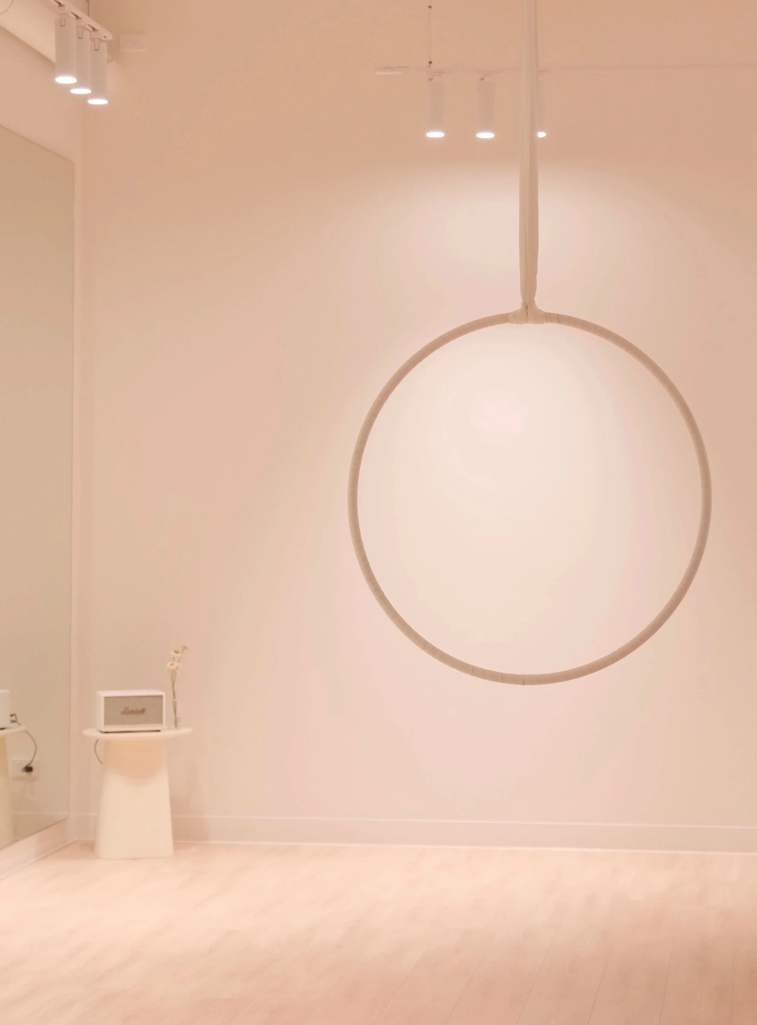 Minimalist interior with a hanging circular yoga hoop, a small table with a speaker and decorative branch on top, and a large mirror on the far wall.