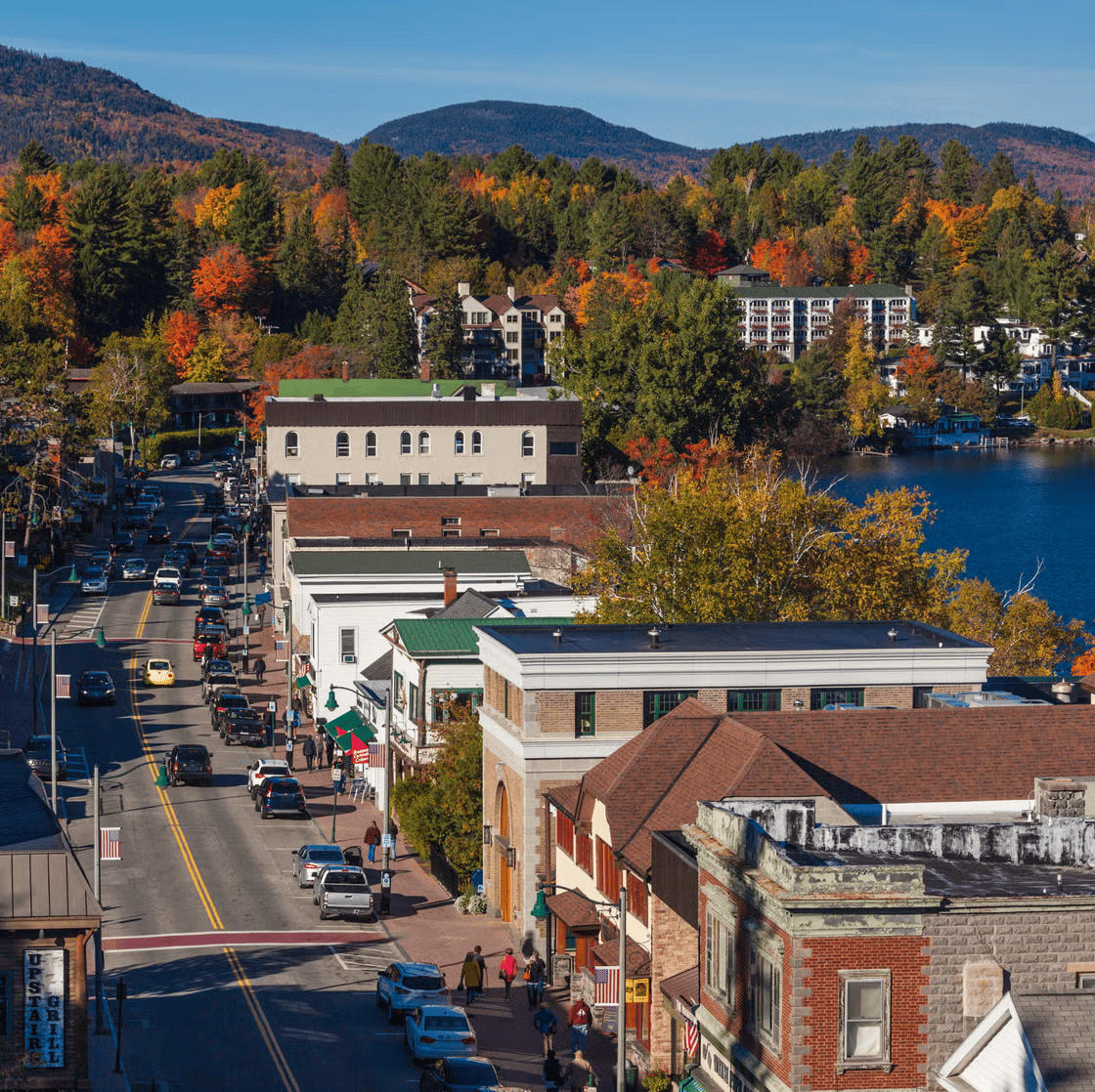 A small town with brick and wooden buildings along a busy street, colorful trees, a lake, and mountains in the background during autumn.