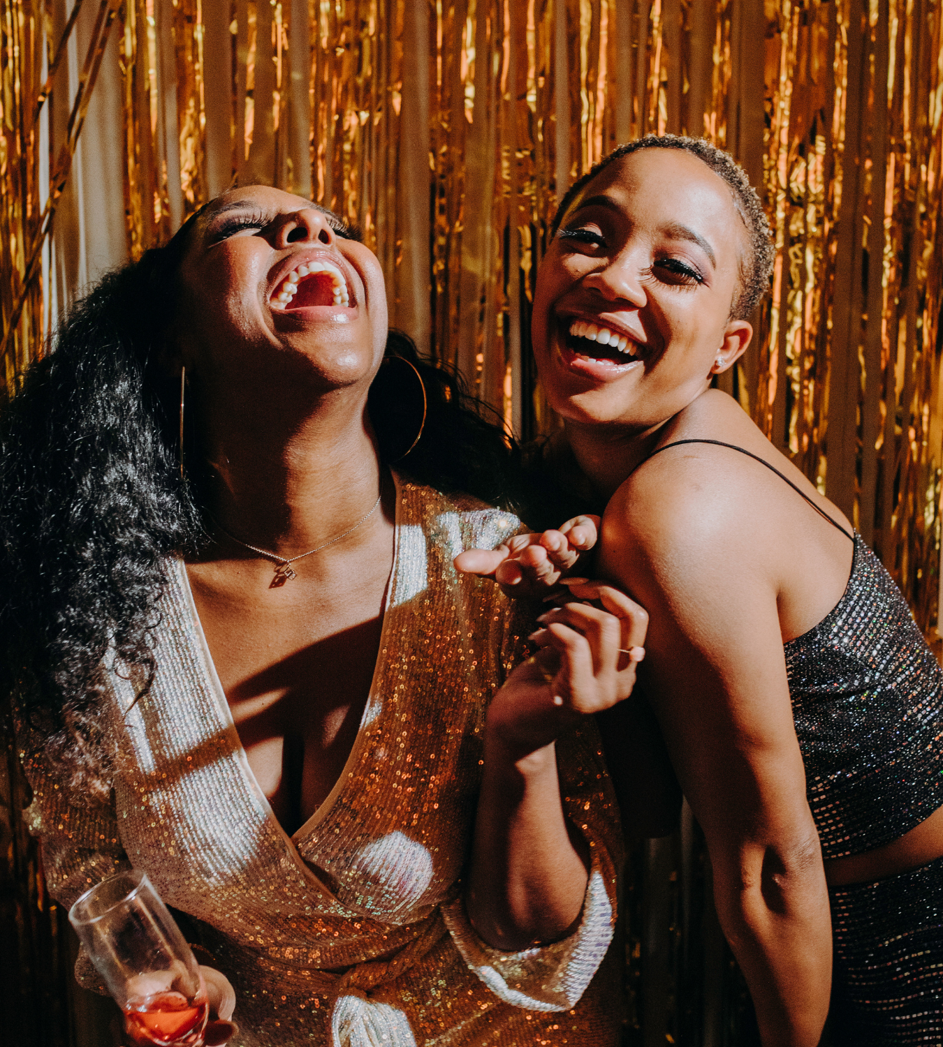 Two women laughing and enjoying a fun moment at a party with a golden, glittery backdrop.
