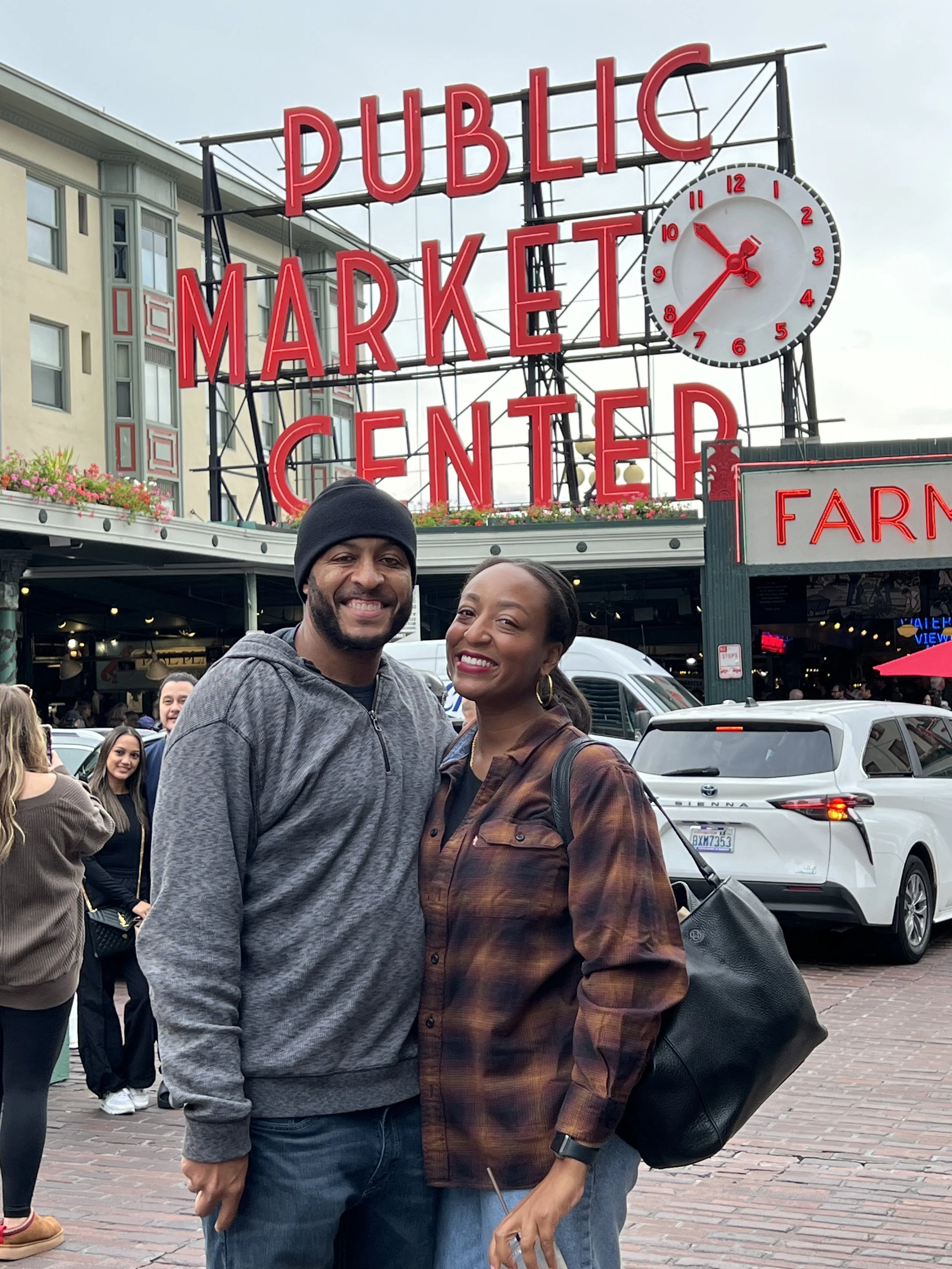 Two smiling people standing in front of the Public Market Center sign, with a clock and parked cars behind them.