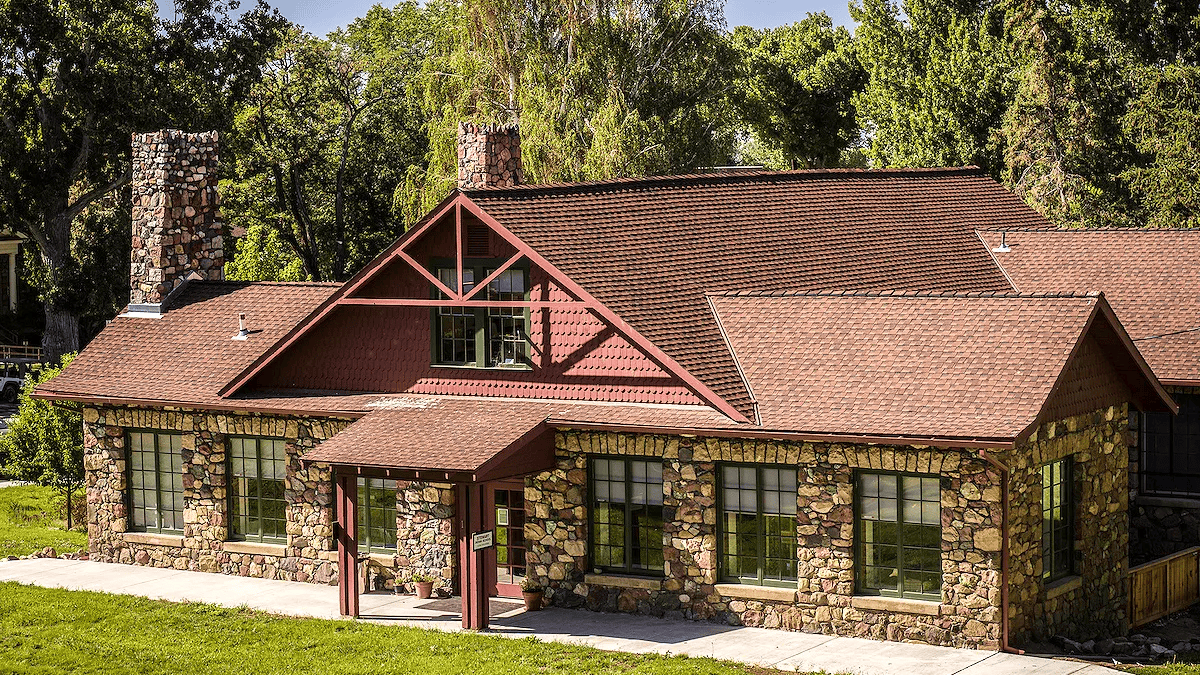 Stone house with large windows, red roof, and chimney, surrounded by trees.