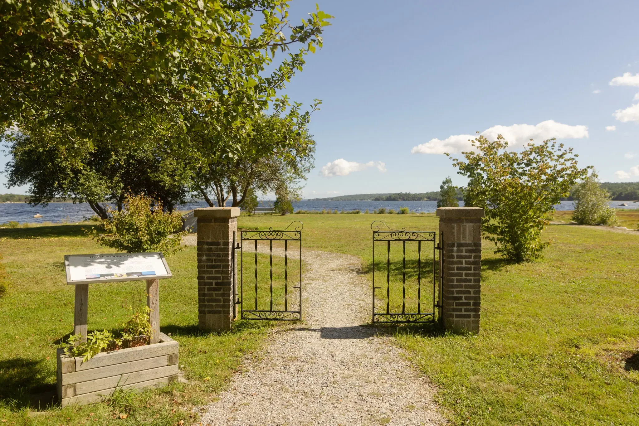 View of an open wrought-iron gate leading to a grassy riverbank with trees, a lake, and a partly cloudy sky in the background.