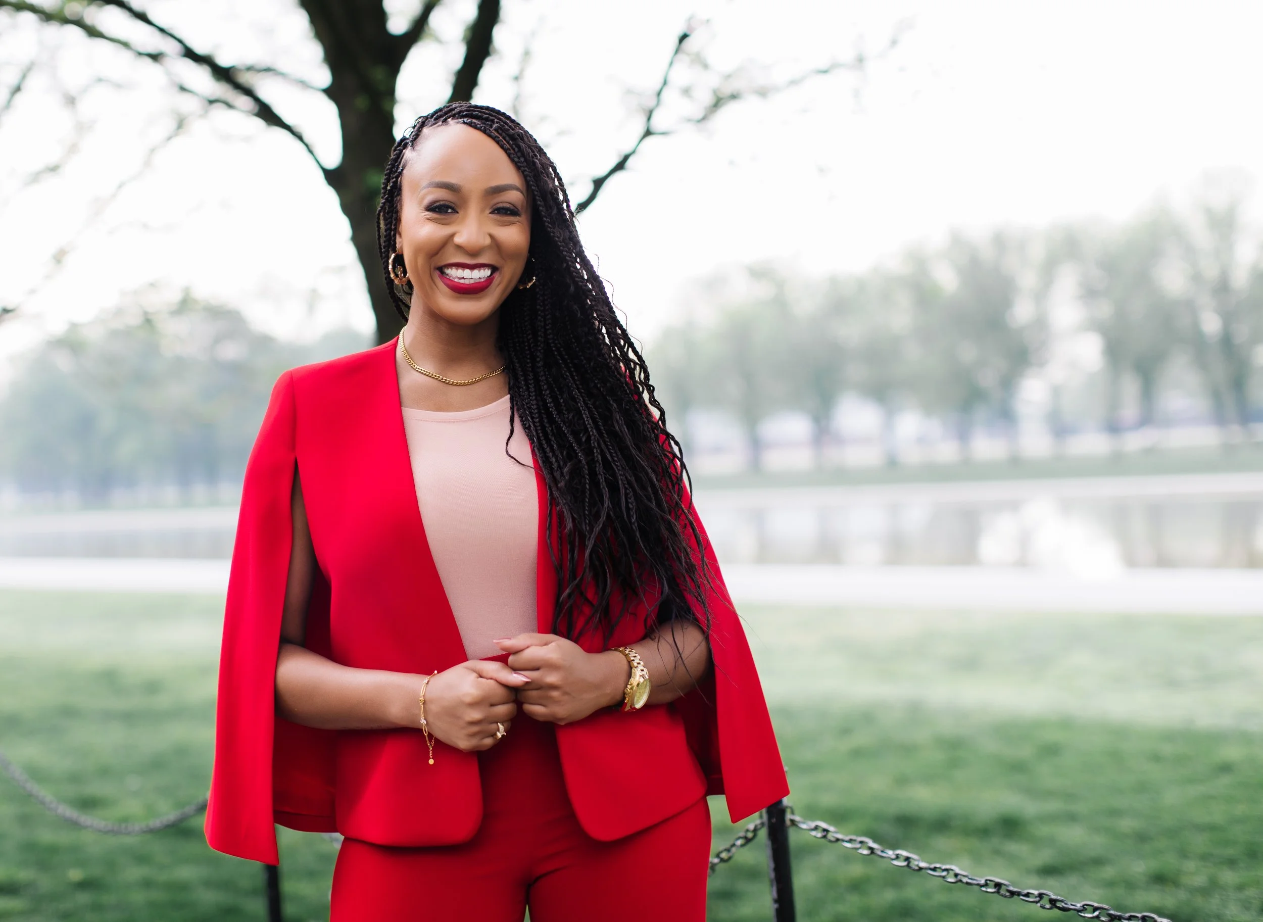 A woman in a red blazer and red pants standing outdoors near a park with trees and water in the background, smiling and looking at the camera.