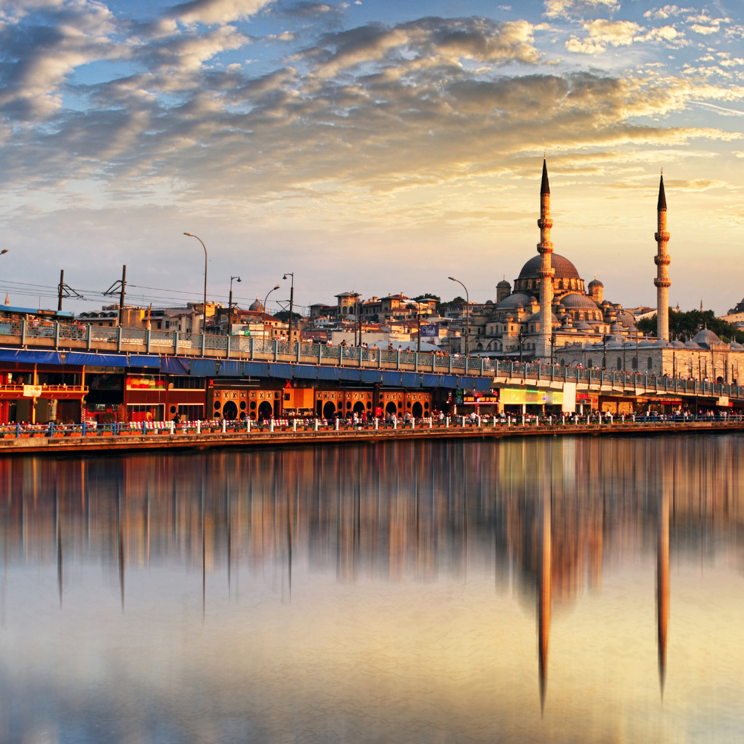 A cityscape during sunset featuring a mosque with two tall minarets, a bridge, and reflections on the water.