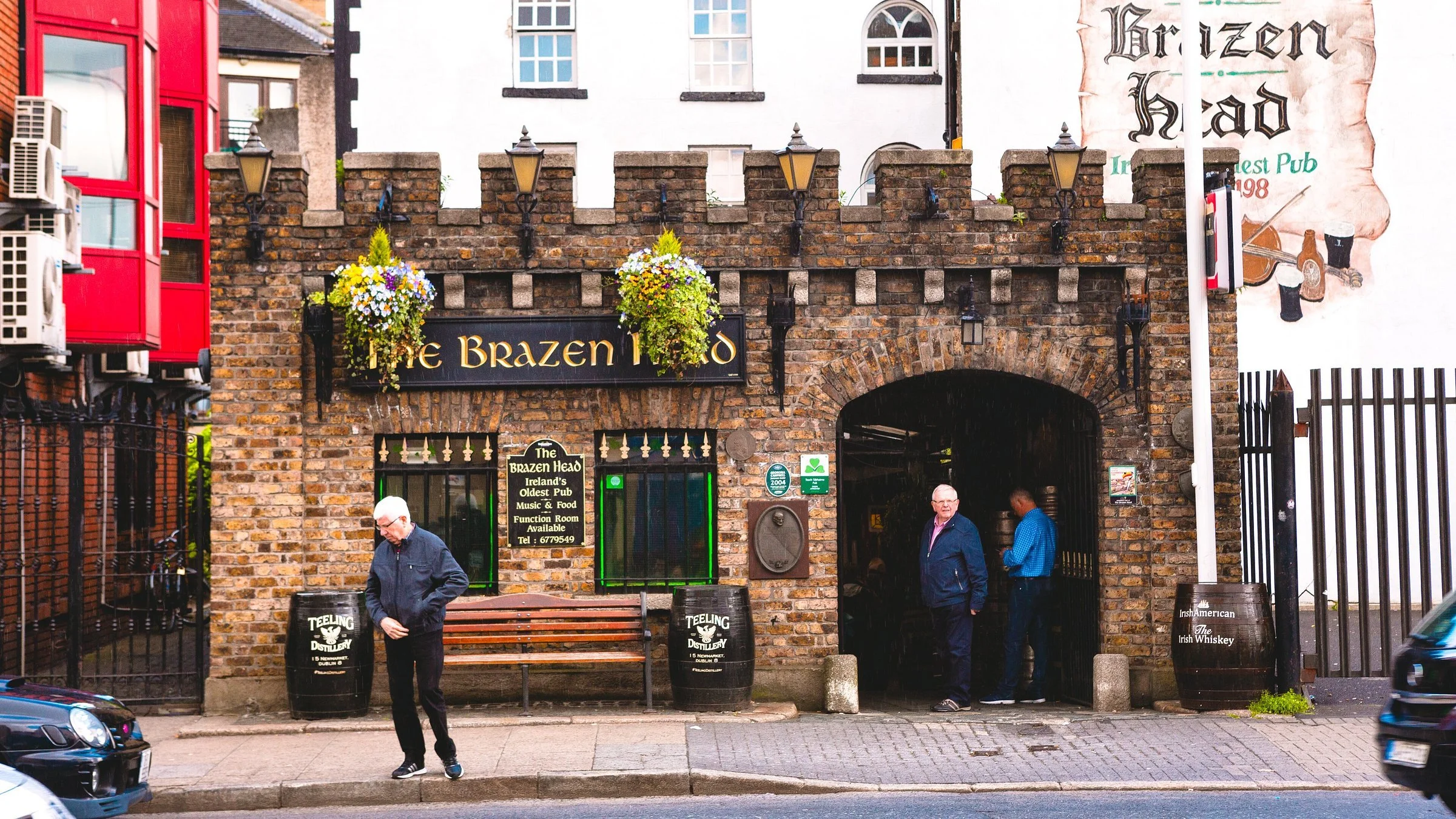 The exterior of The Brazen Head pub in Dublin, Ireland, featuring a brick archway entrance, hanging flower baskets, and signage indicating it's Ireland's oldest pub. There are a few people near the entrance and two barrels outside.