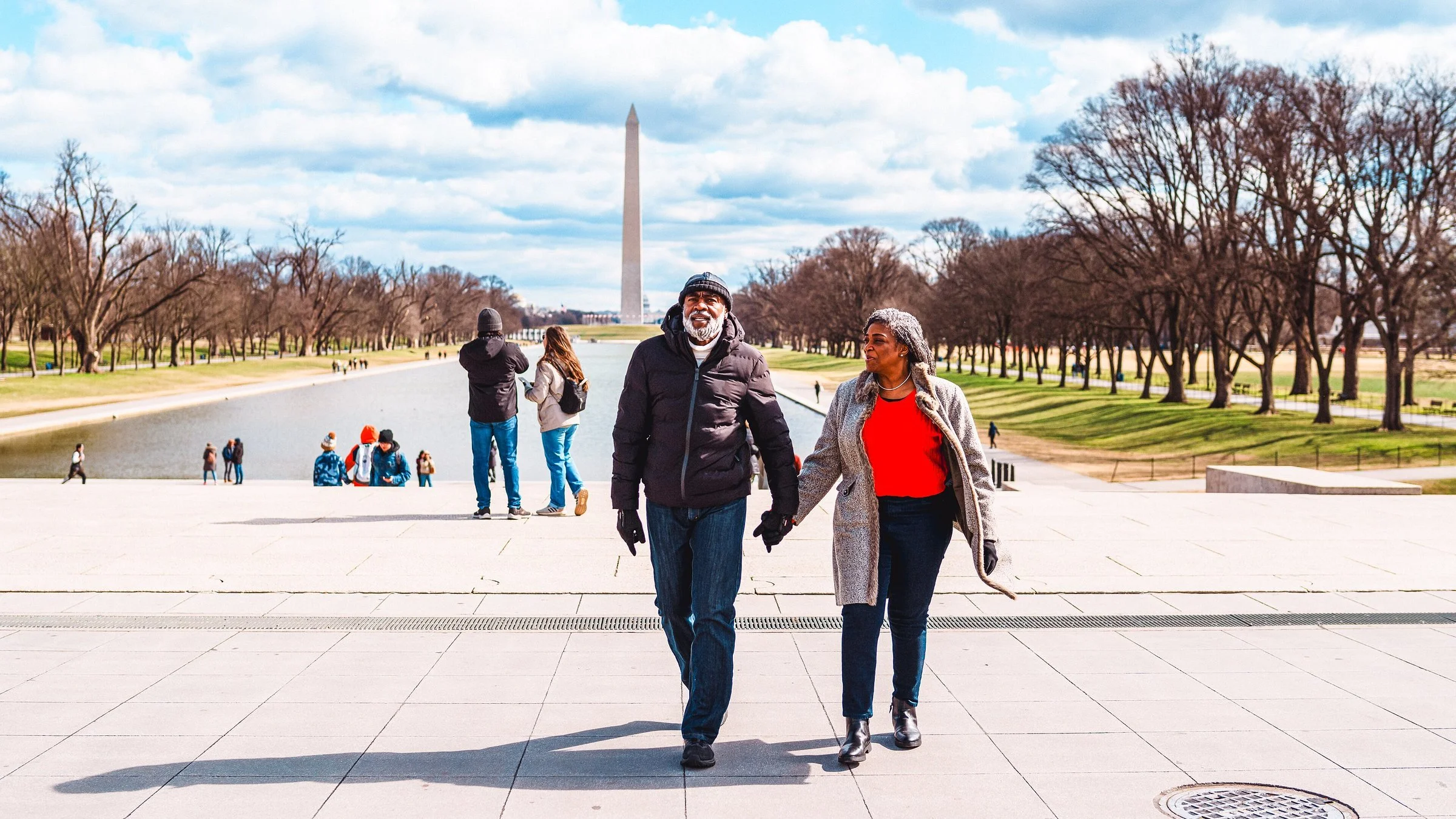 A couple walks hand in hand along the National Mall with the Washington Monument in the background on a sunny day with few clouds.