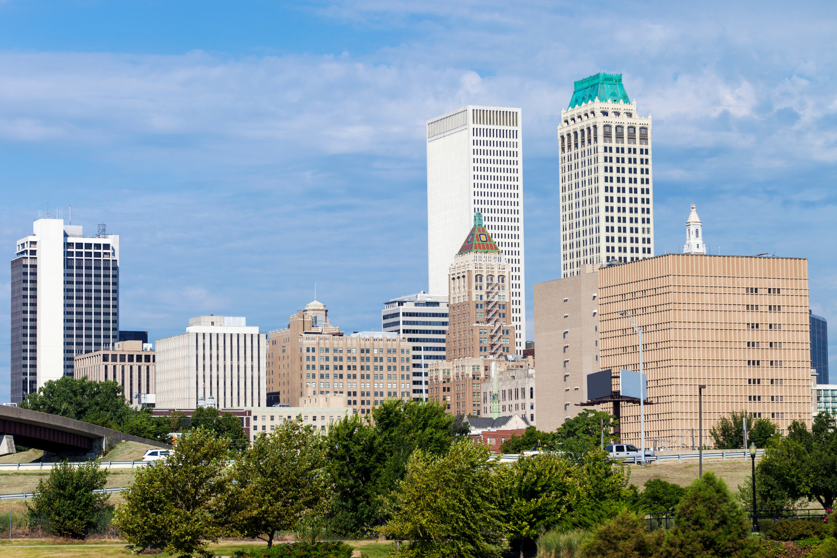Skyline of a city with tall skyscrapers, some with distinctive architectural features, topped with a partly cloudy blue sky and lush green trees in the foreground.