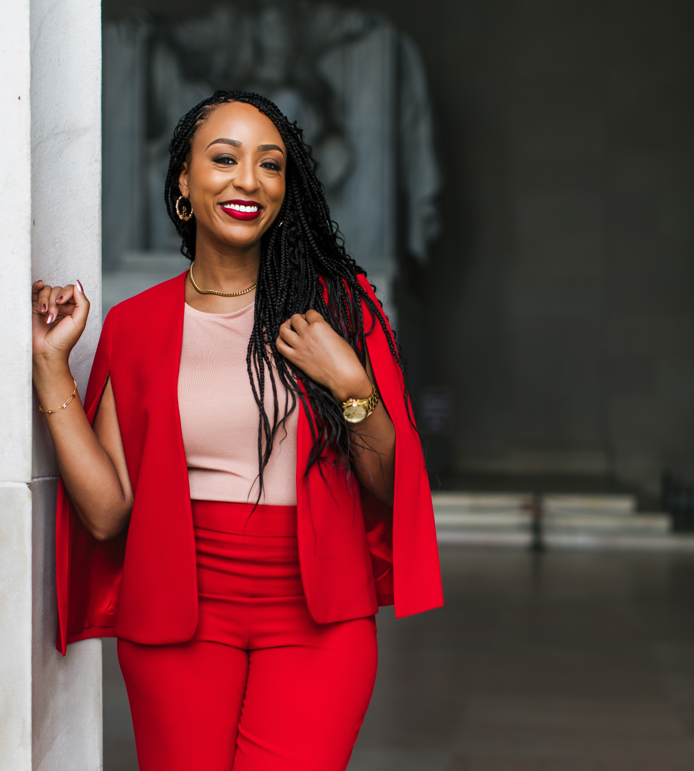 A woman with long black braids, wearing a red blazer and matching red pants, standing and smiling while leaning against a column.