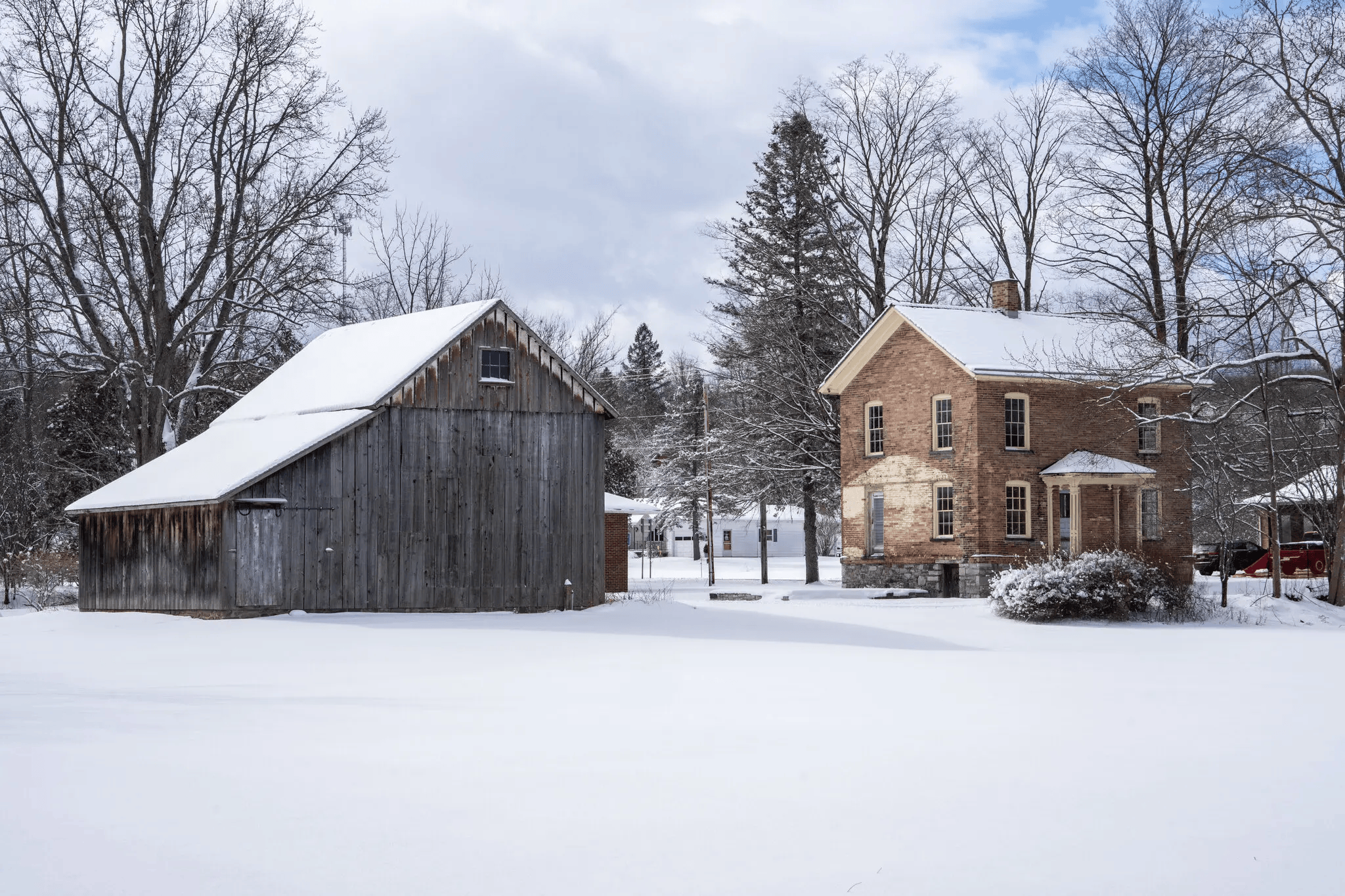 A snowy scene featuring a wooden barn on the left and a brick house on the right, surrounded by leafless trees under cloudy sky.