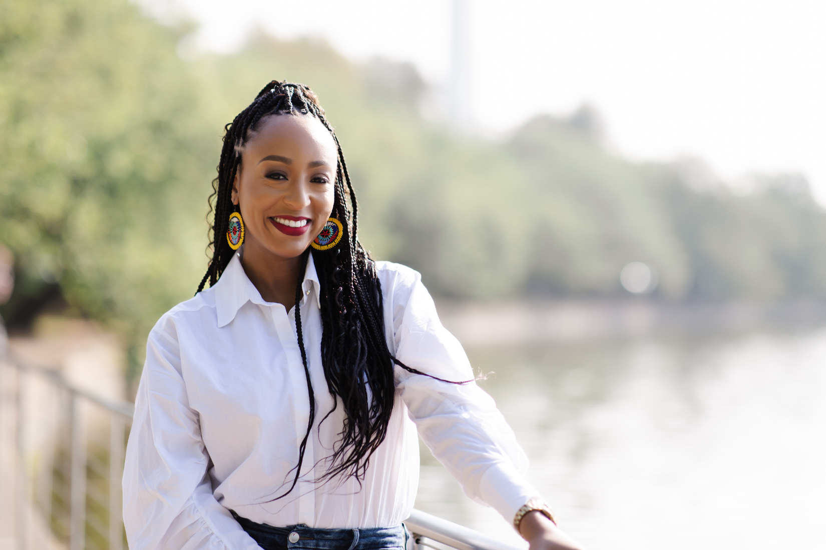 A smiling woman with long braids, wearing colorful beaded earrings, a white button-up shirt, and blue jeans, standing outdoors near a body of water with trees in the background.