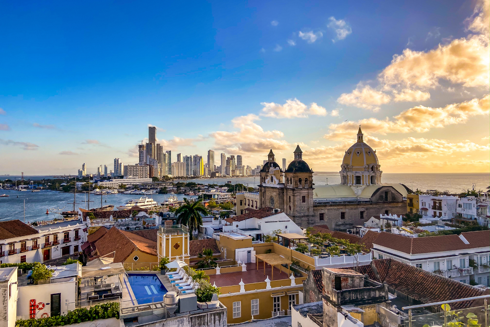 Sunset over a city skyline with high-rise buildings, a harbor with boats, and historic architecture including a church with a yellow dome, in a coastal urban area.
