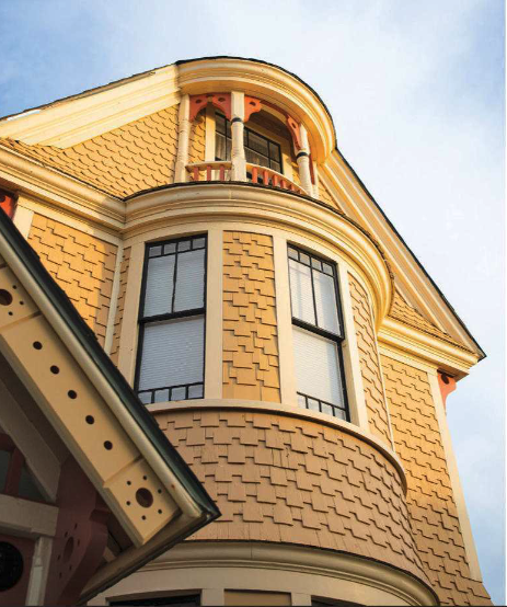 Close-up of a yellow Victorian-style house with decorative shingles, a small balcony, and tall windows, with part of the roof and sky visible.
