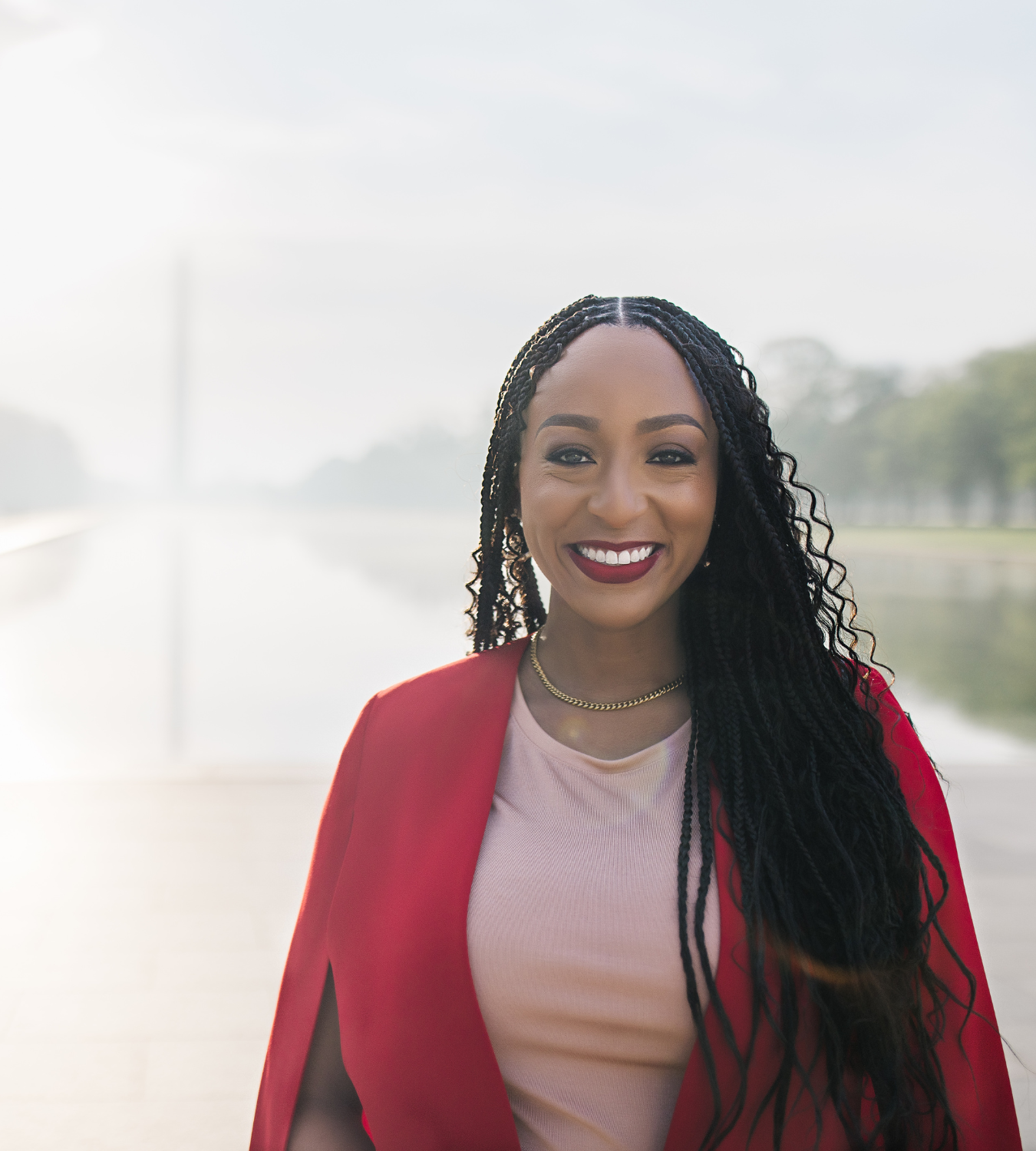 Smiling woman with long curly black hair, wearing a beige top, red blazer, gold necklace, outdoors near a body of water, bright sky.