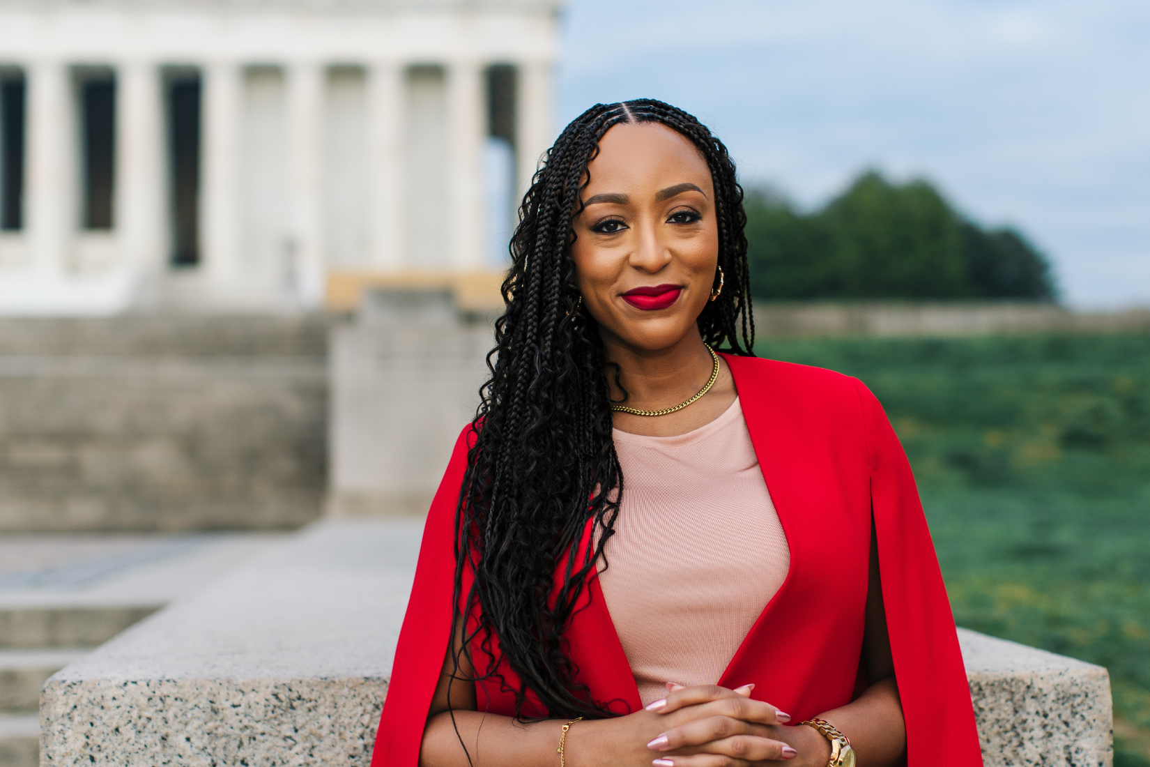 A woman with long braided hair wearing a red sleeveless blazer and a beige top, standing outdoors near a stone railing with a historic building and green landscape in the background.