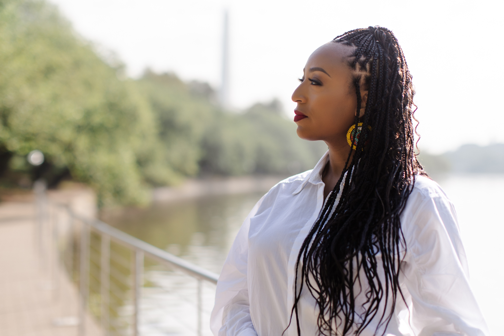 A woman with braided hair and earrings looking to the side outdoors near a body of water with trees in the background.