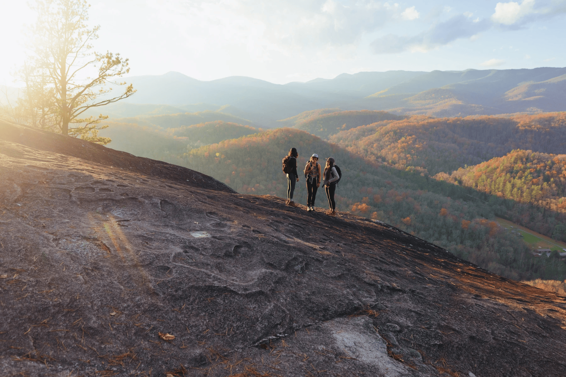 Three hikers with backpacks standing on a rocky mountain overlook at sunset, overlooking rolling hills and mountains covered in fall foliage.