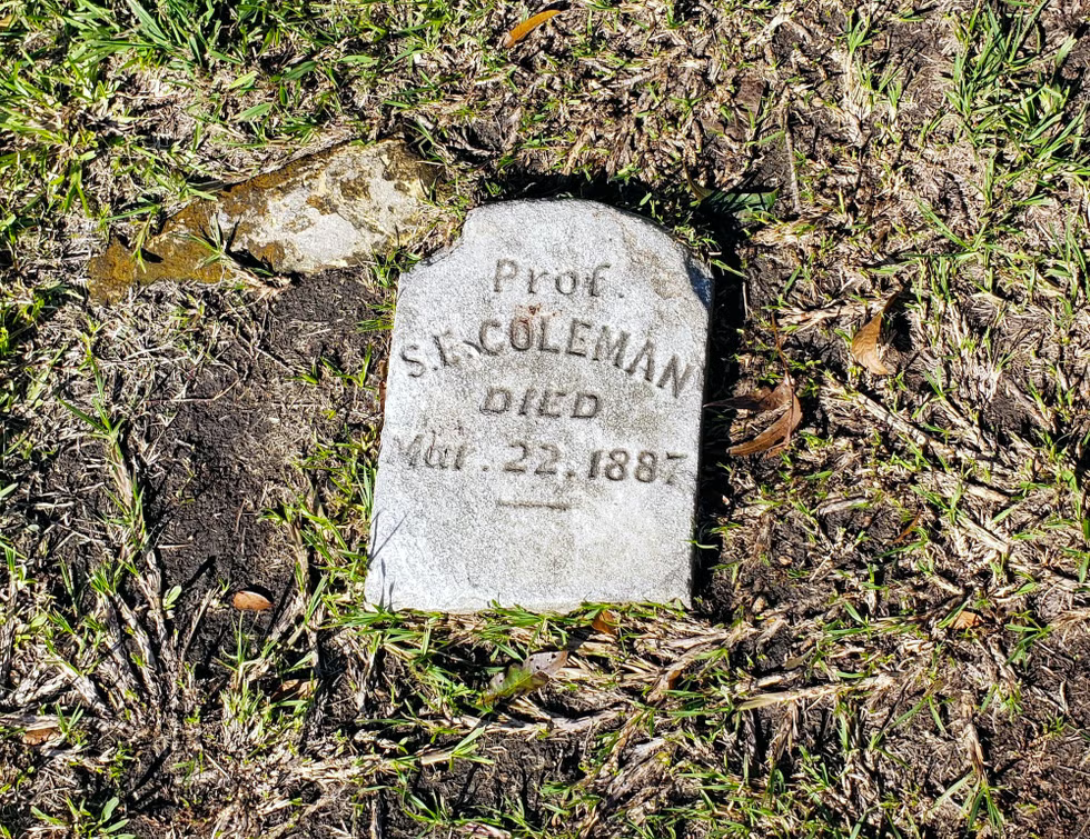 A small, weathered grave marker on the ground. The headstone reads: 'Prof. S. C. Coleman Died March 22, 1887.' Surrounding grass and soil.