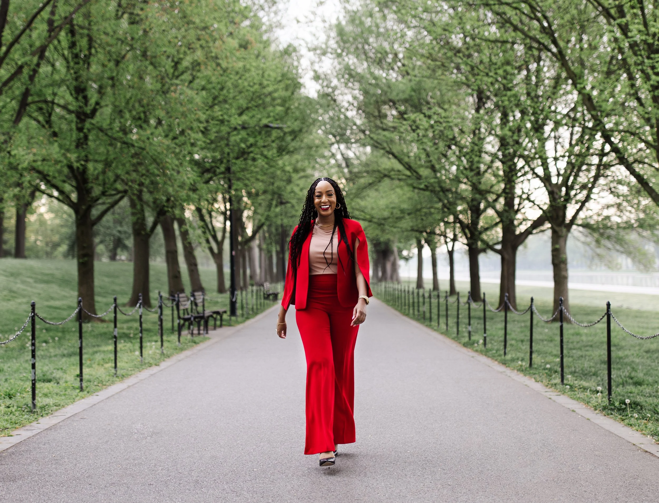 A woman walking down a paved park path lined with green trees, wearing a red suit and smiling.