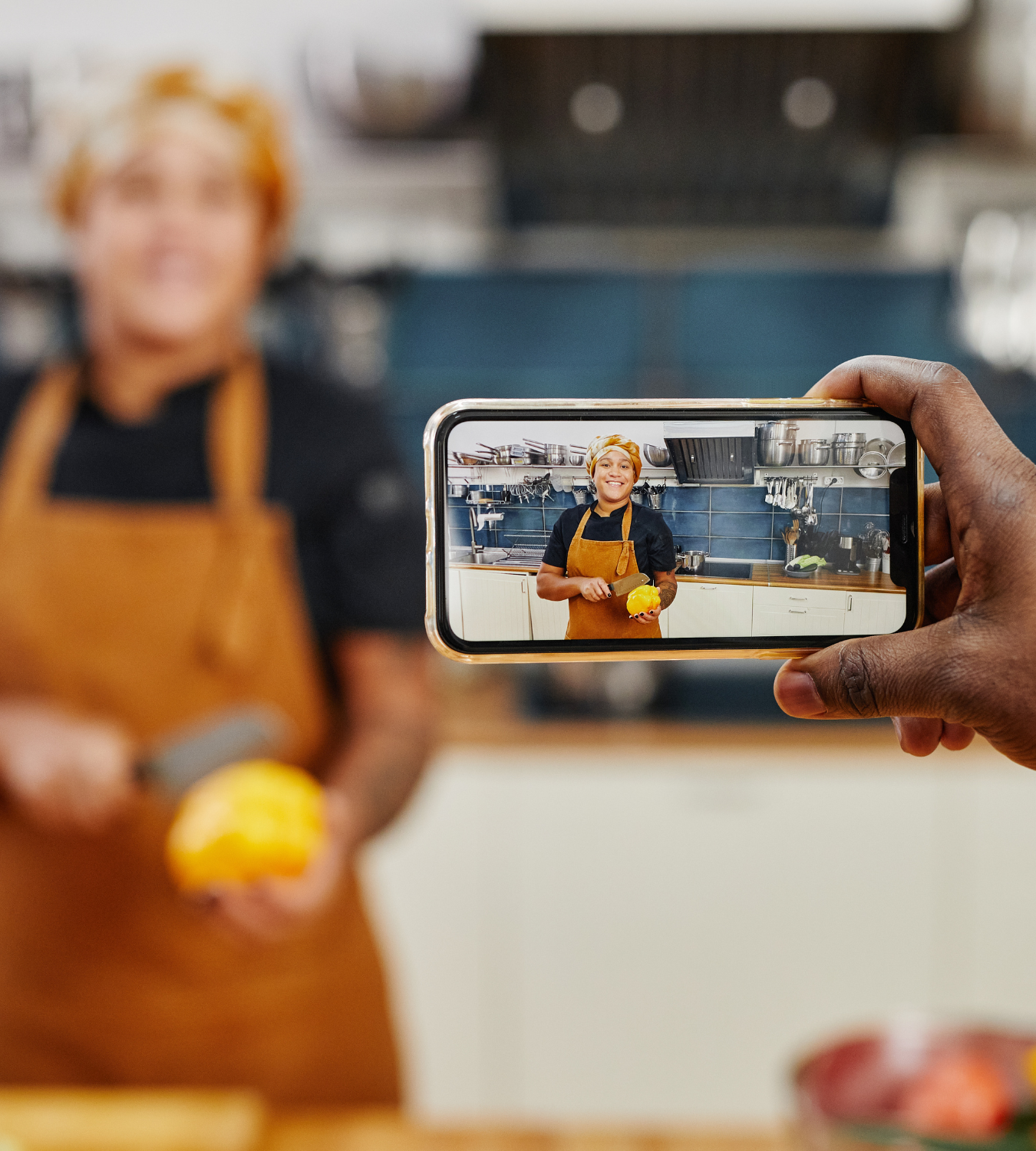 Person holding a phone, taking a photo of a smiling woman in a kitchen holding a lemon, with kitchen appliances in the background.