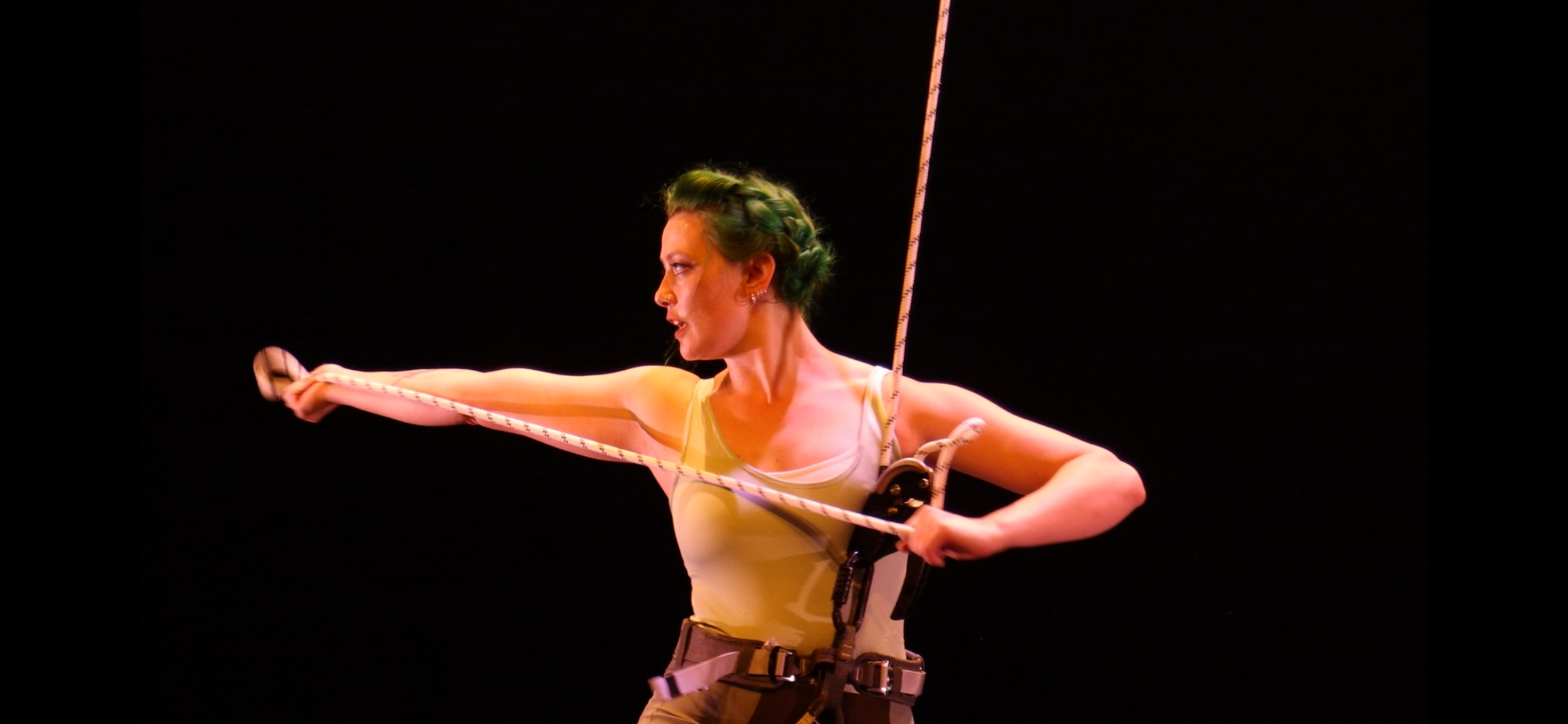 A woman with green hair wearing a beige tank top, holding the end of the rope that she is performing low flow aerial dance in harness on, standing against a dark background.