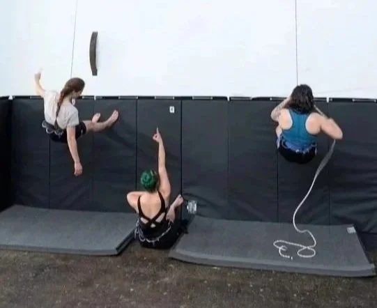 Three women practicing vertical dance on a white indoor wall, with one woman sitting on the floor to demonstrate a move and two women climbing.