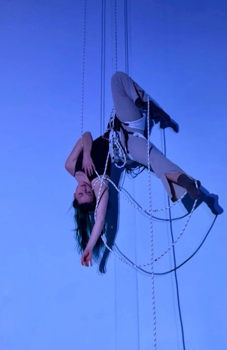 Emily with long hair hanging upside down, performing vertical dance, surrounded by ropes, posing against a blue background.