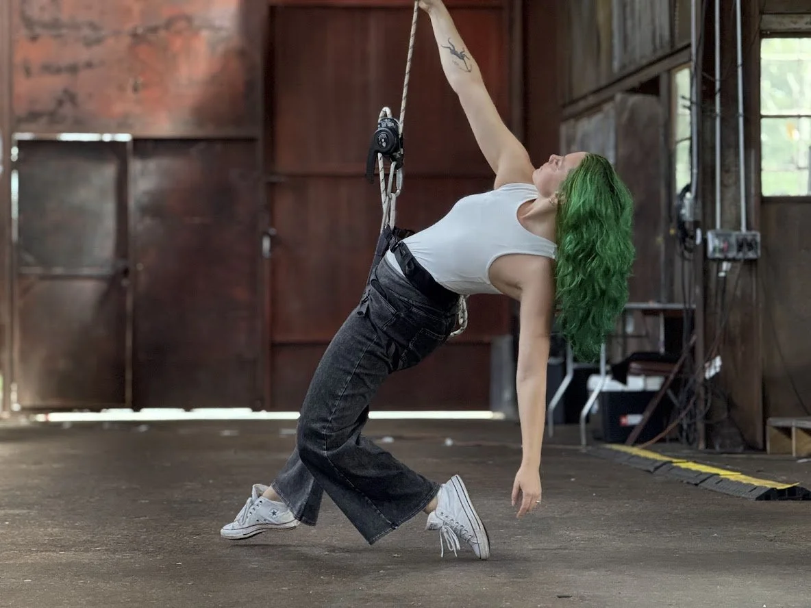 Emily with green hair performing a dance move in harness, wearing a white sleeveless top, black pants, and white sneakers, inside a warehouse