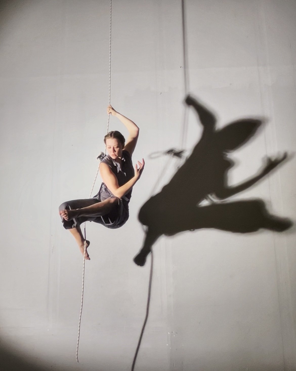 A woman performing an aerial wall dance act on a rope, with a shadow cast on the wall behind her.