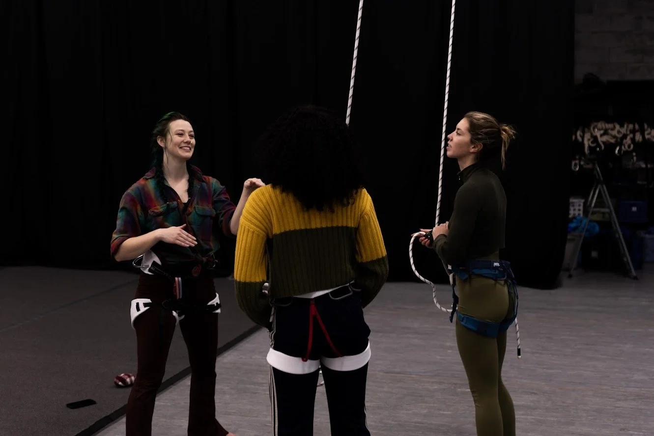 Three women standing in a dark indoor climbing gym, dressed in climbing harnesses , preparing for harness dance