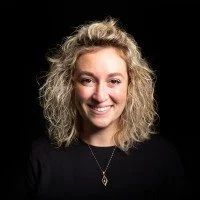A woman with curly blonde hair smiling, wearing a black top and a necklace.