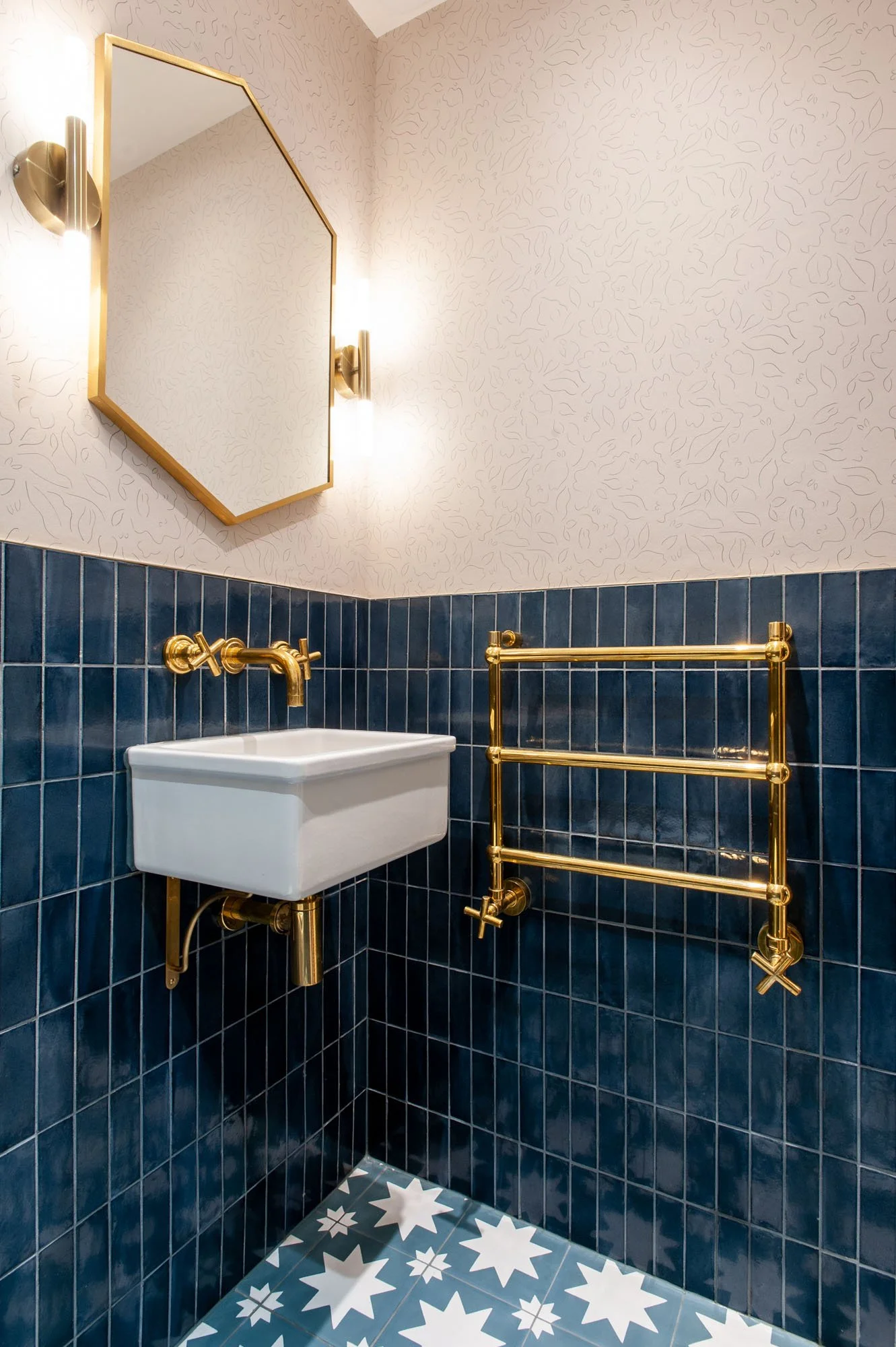 A bathroom with dark blue tiled walls, a small white sink with gold fixtures, a gold towel rack, a mirror with a gold frame, and geometric patterned floor tiles.