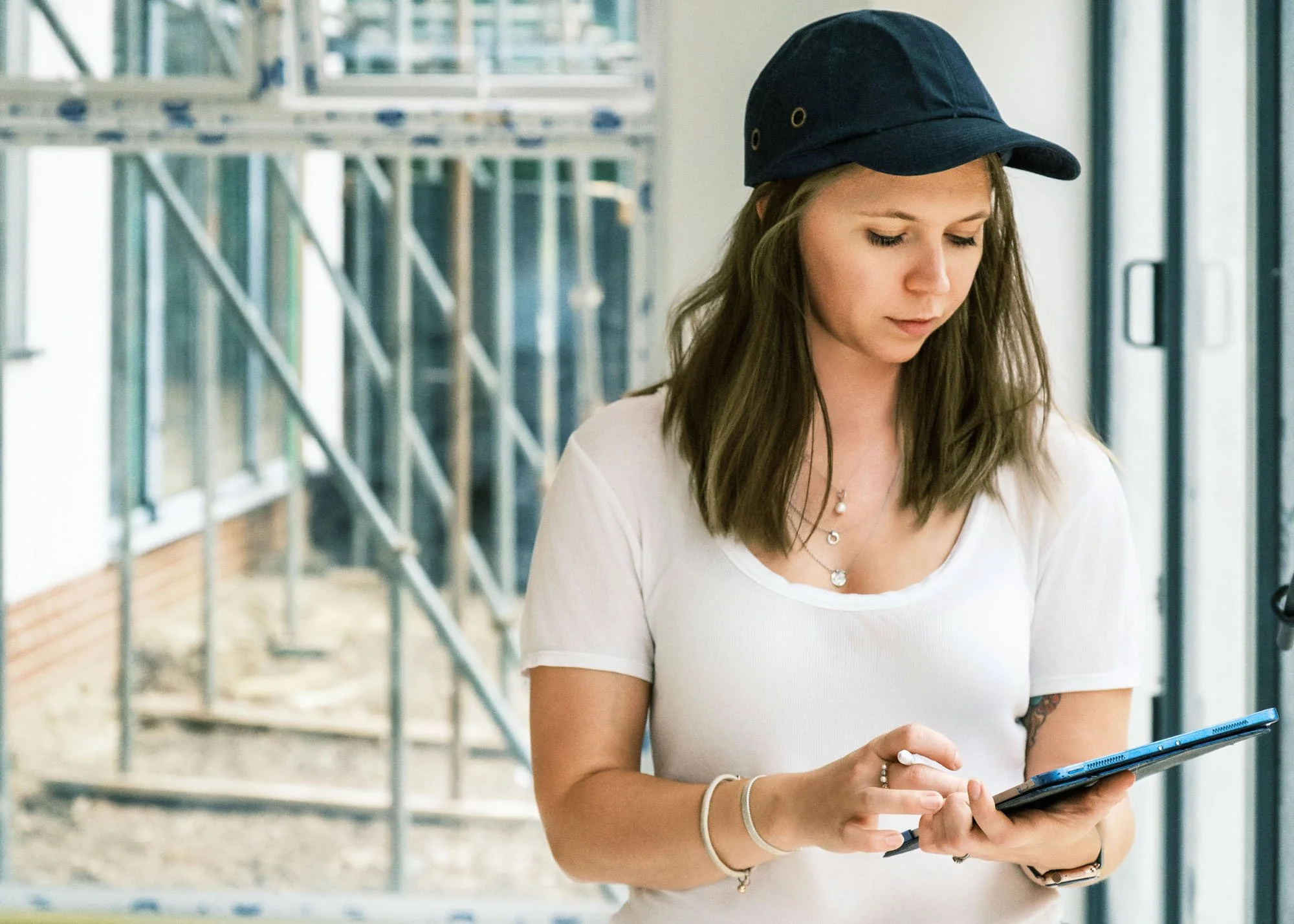 A young woman with shoulder-length brown hair, wearing a white t-shirt and a navy blue baseball cap, is standing indoors near large windows, looking at and using a tablet or phone.