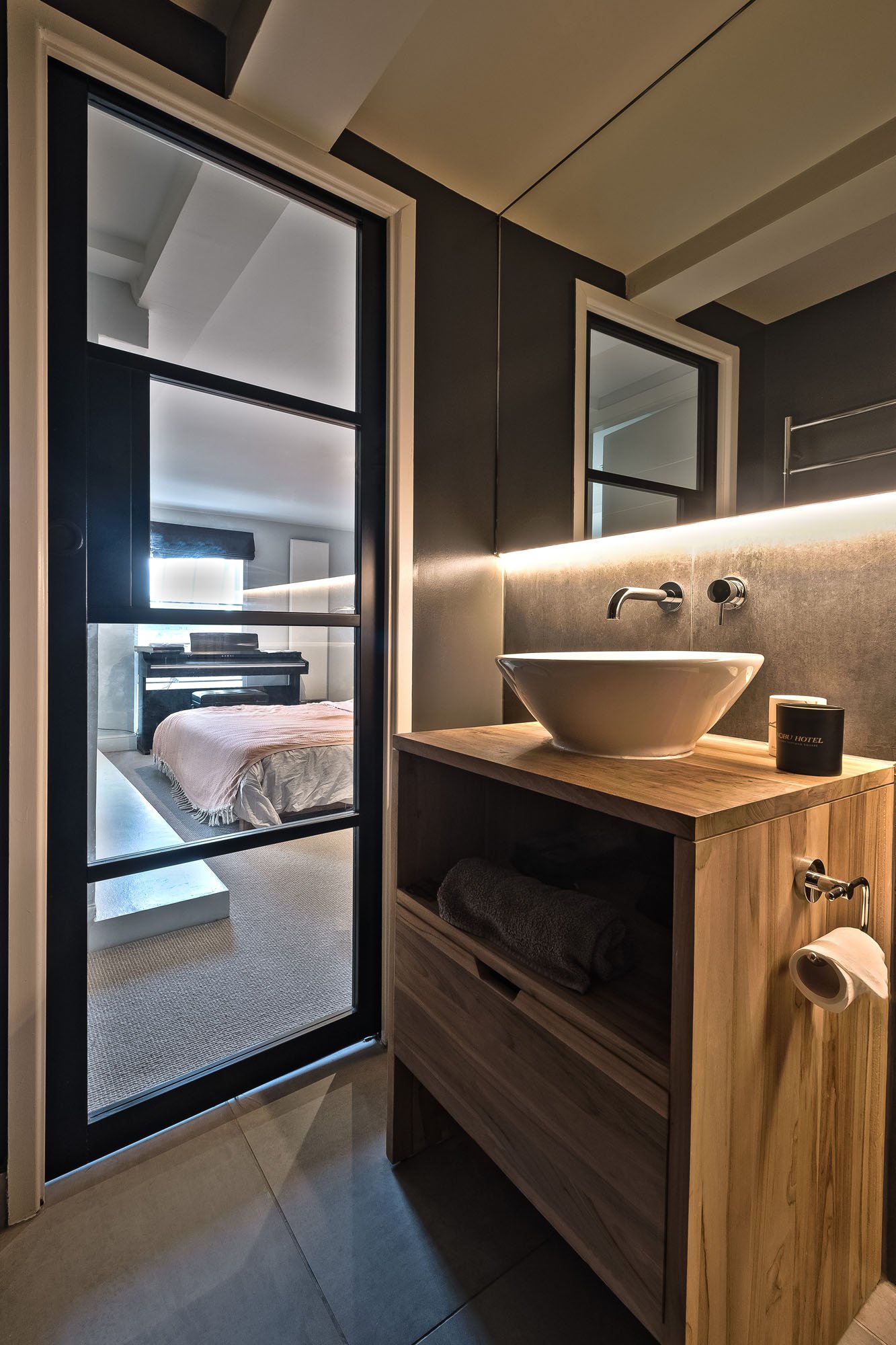 A modern bathroom with a wooden vanity featuring a vessel sink, wall-mounted faucet, and a mirror with LED lighting. Through a glass door, a bedroom with a bed and a dresser is visible.