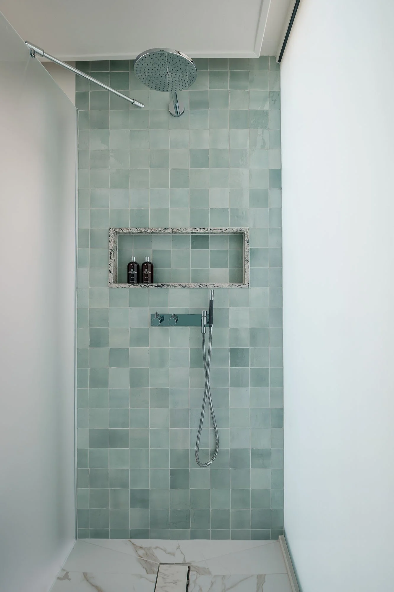 Minimalist shower with large rain shower head, built-in shelf with two bottles, and handheld shower head, in a bathroom with light green tiles.