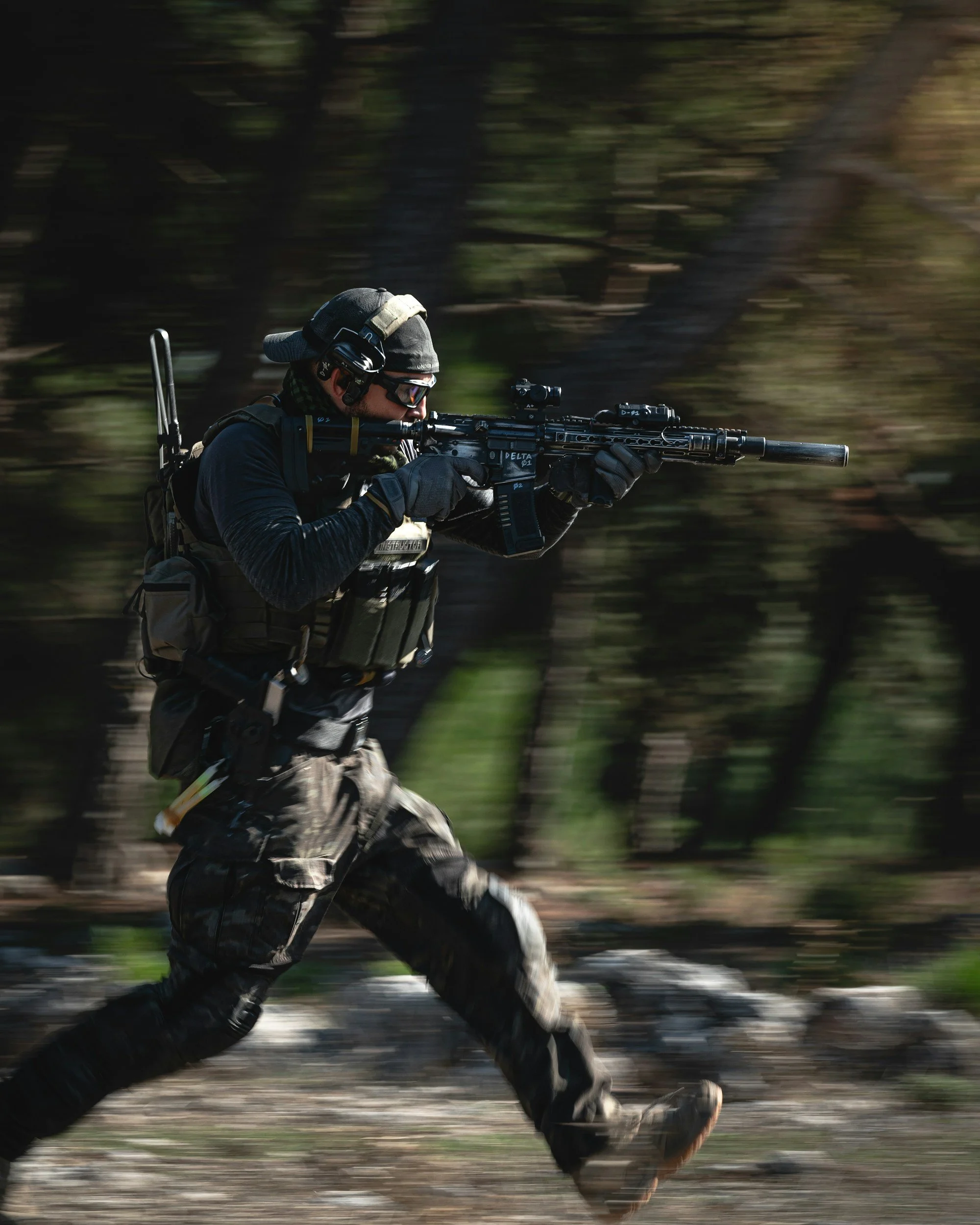 A soldier running outdoors holding a scoped rifle, with a blurred background of trees and rocks.