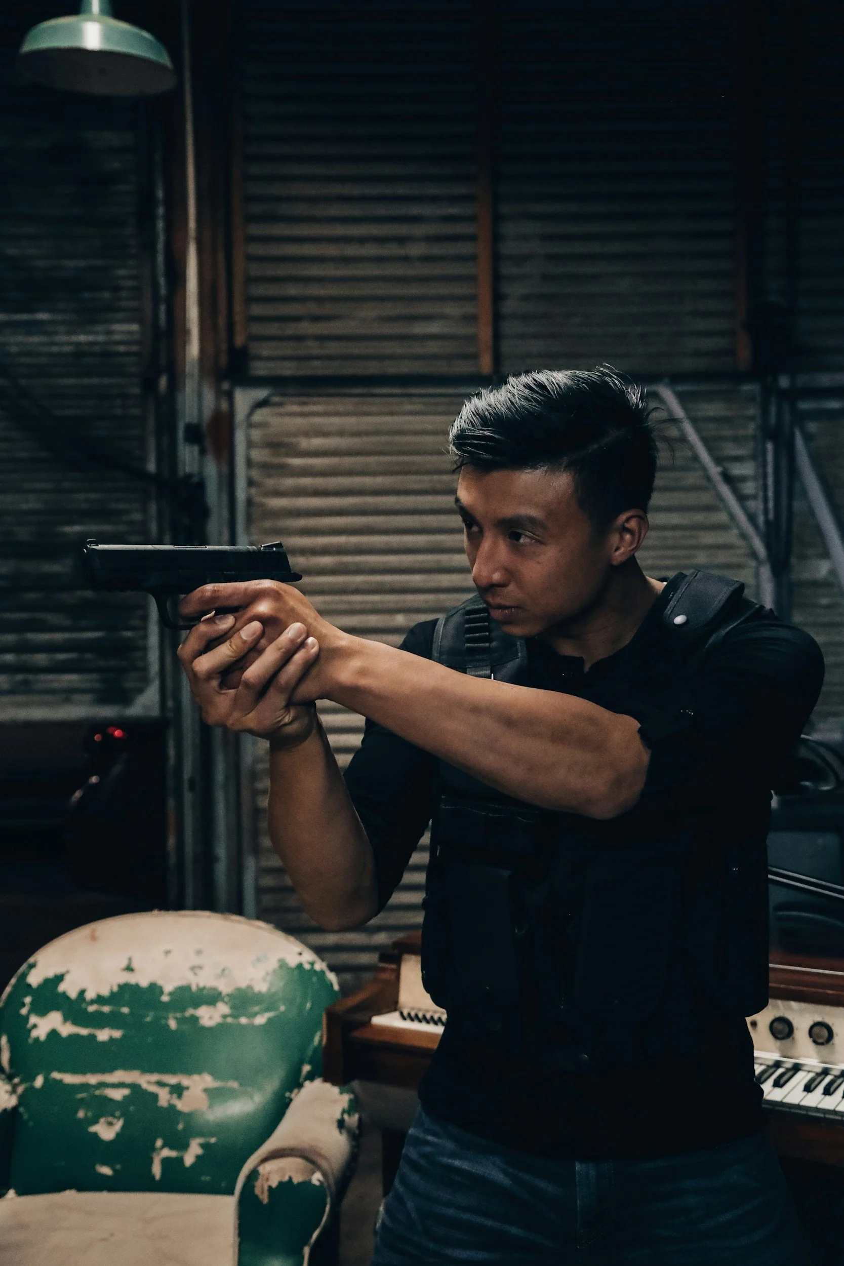 A man in black attire aiming a handgun indoors, with wooden shutters and an old green armchair in the background.