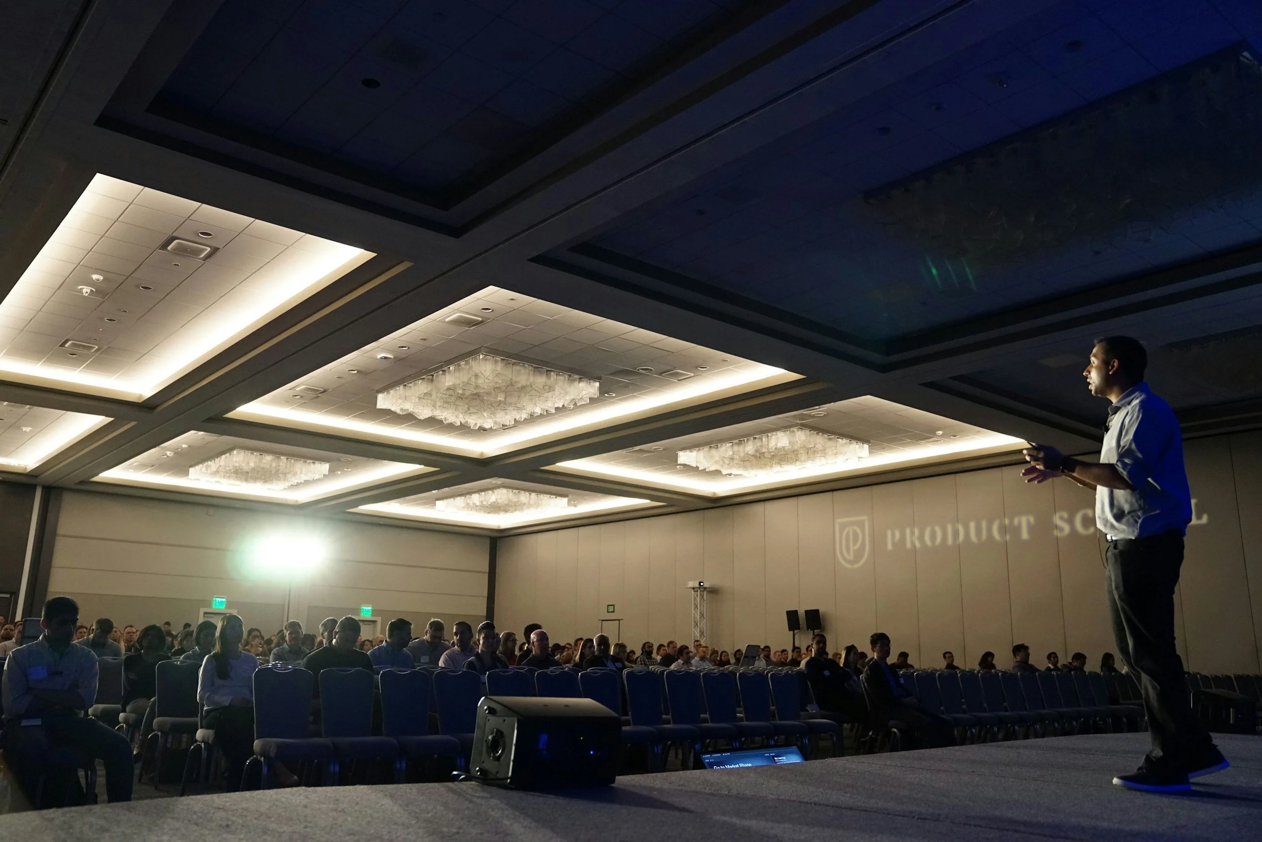 A man giving a presentation on stage at a conference, with a large audience seated in front. The room has bright ceiling lights and a projection on the wall that reads "PRODUCT SCHOOL."