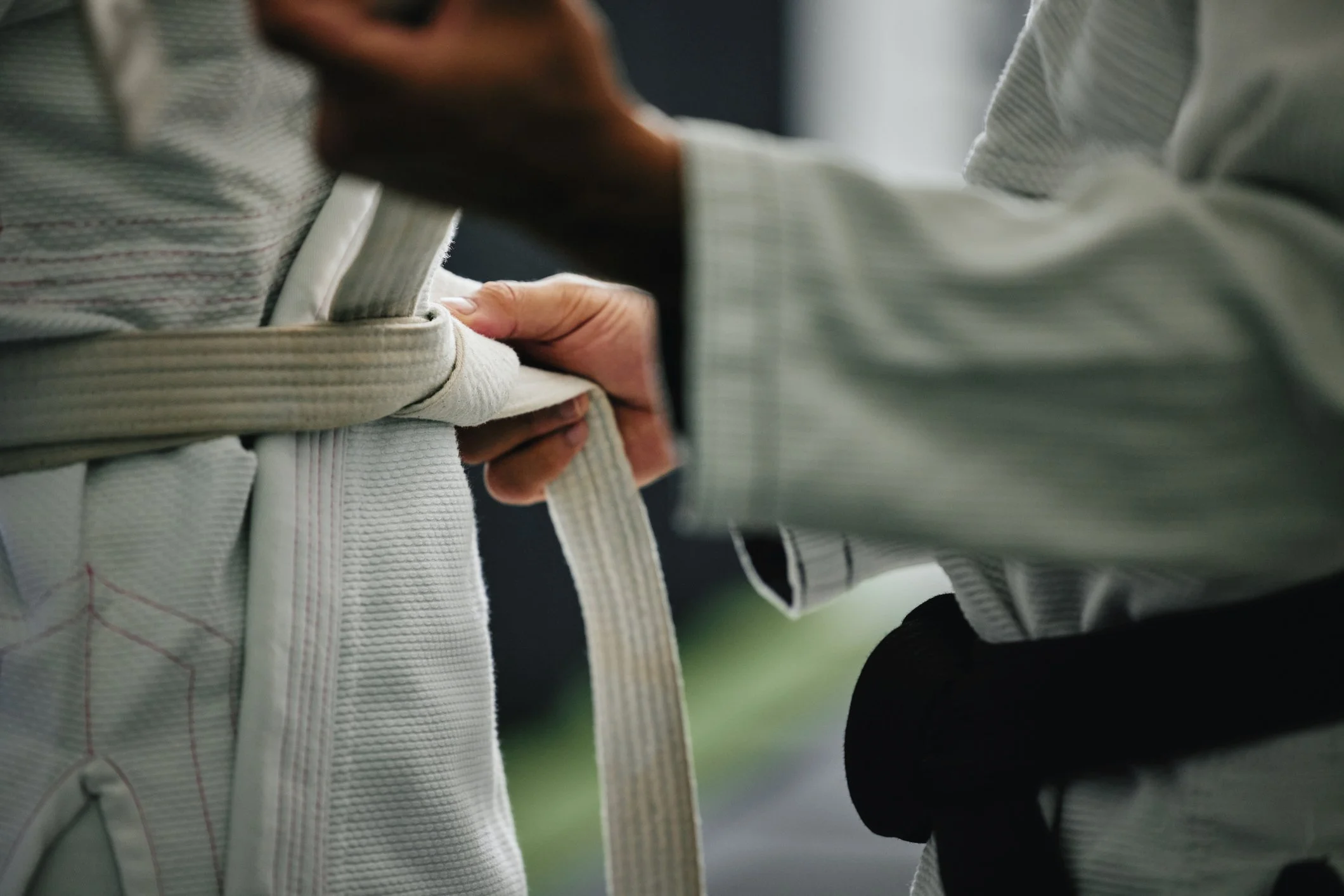 A person tying a white martial arts gi belt around the waist of another individual while dressed in traditional martial arts uniform.