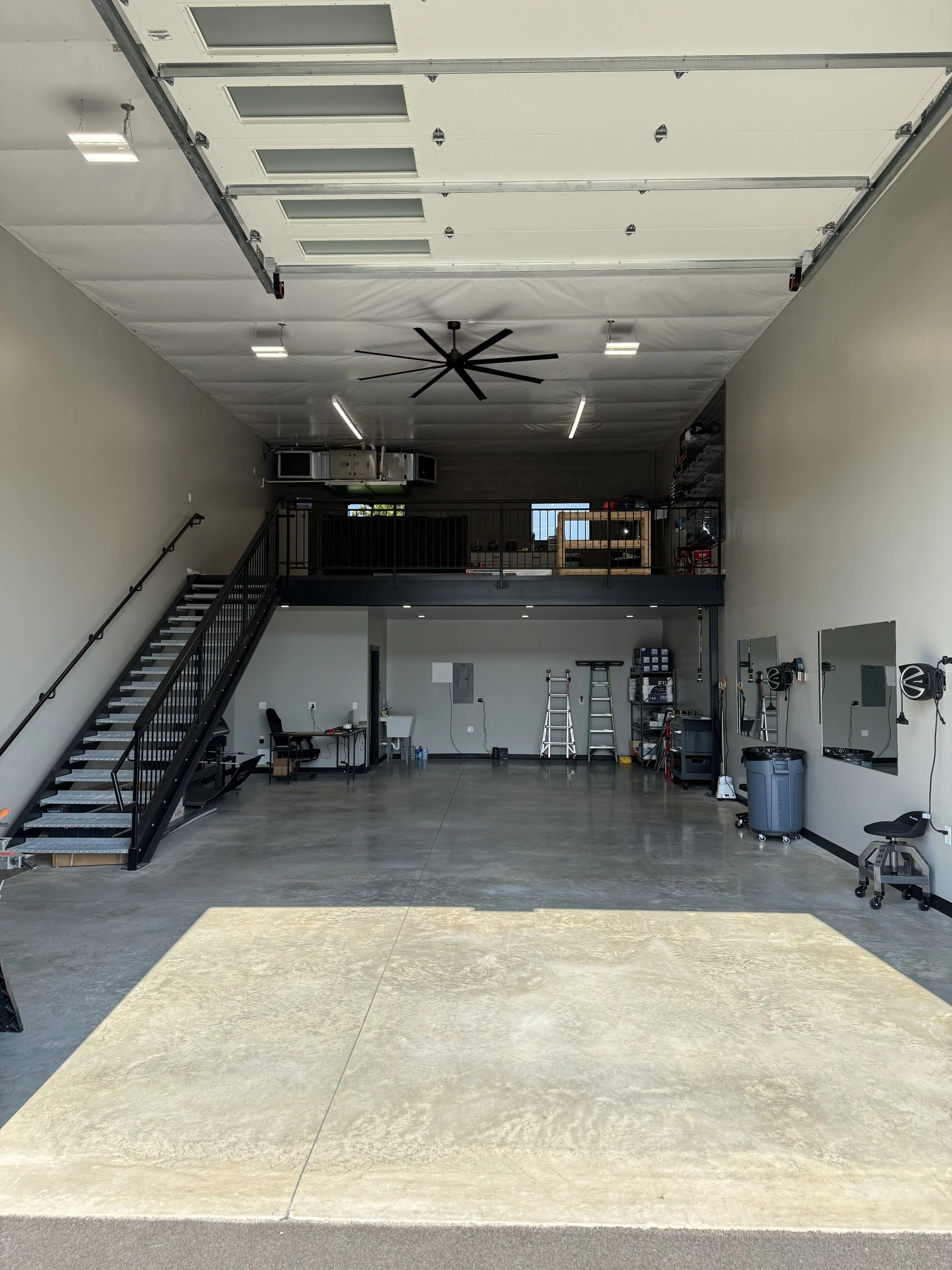 Empty modern garage with high ceiling, large open floor space, laundry sink, stairs leading to a mezzanine, and maintenance tools.