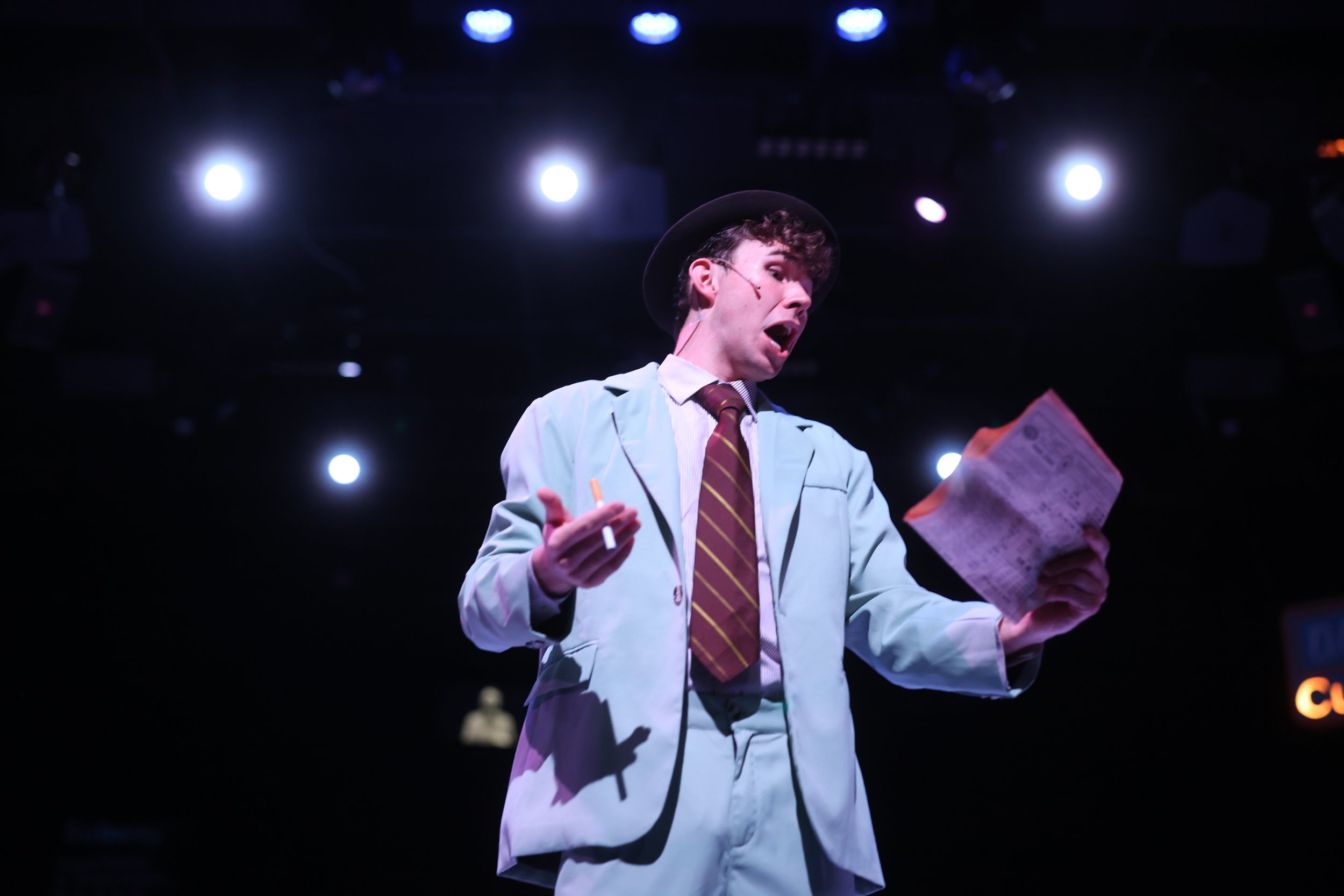 Weller in Guys and Dolls, wearing a light green suit, a dark brown hat, and a striped tie, holding a newspaper in one hand and a cigarette in the other, with stage lighting overhead.