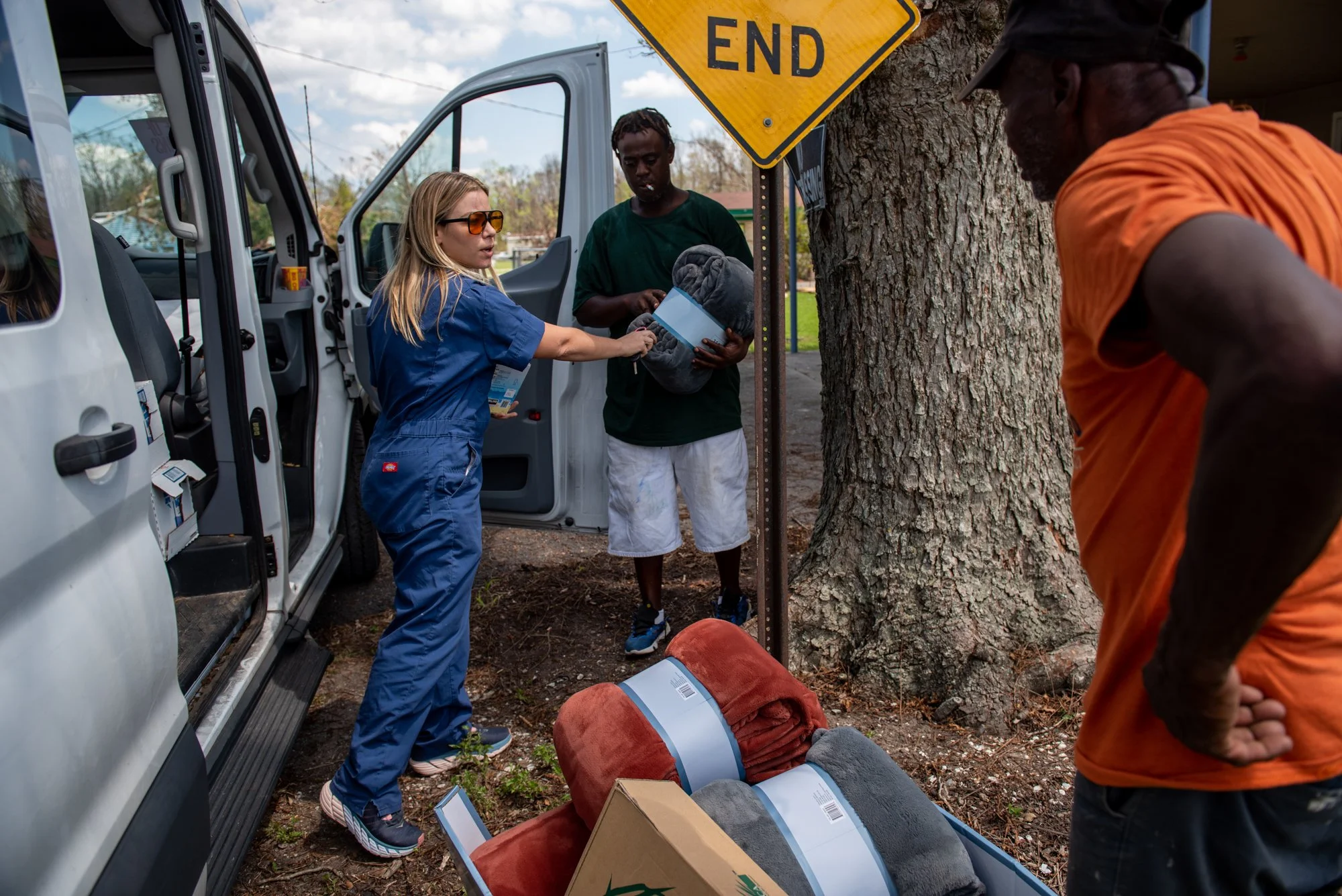 A woman in blue scrubs is handing a blanket to a man in a green shirt and white shorts while another man in an orange shirt looks on. They are outside next to an open white van, with a large tree and an end road sign nearby. There are blankets and boxes on the ground.