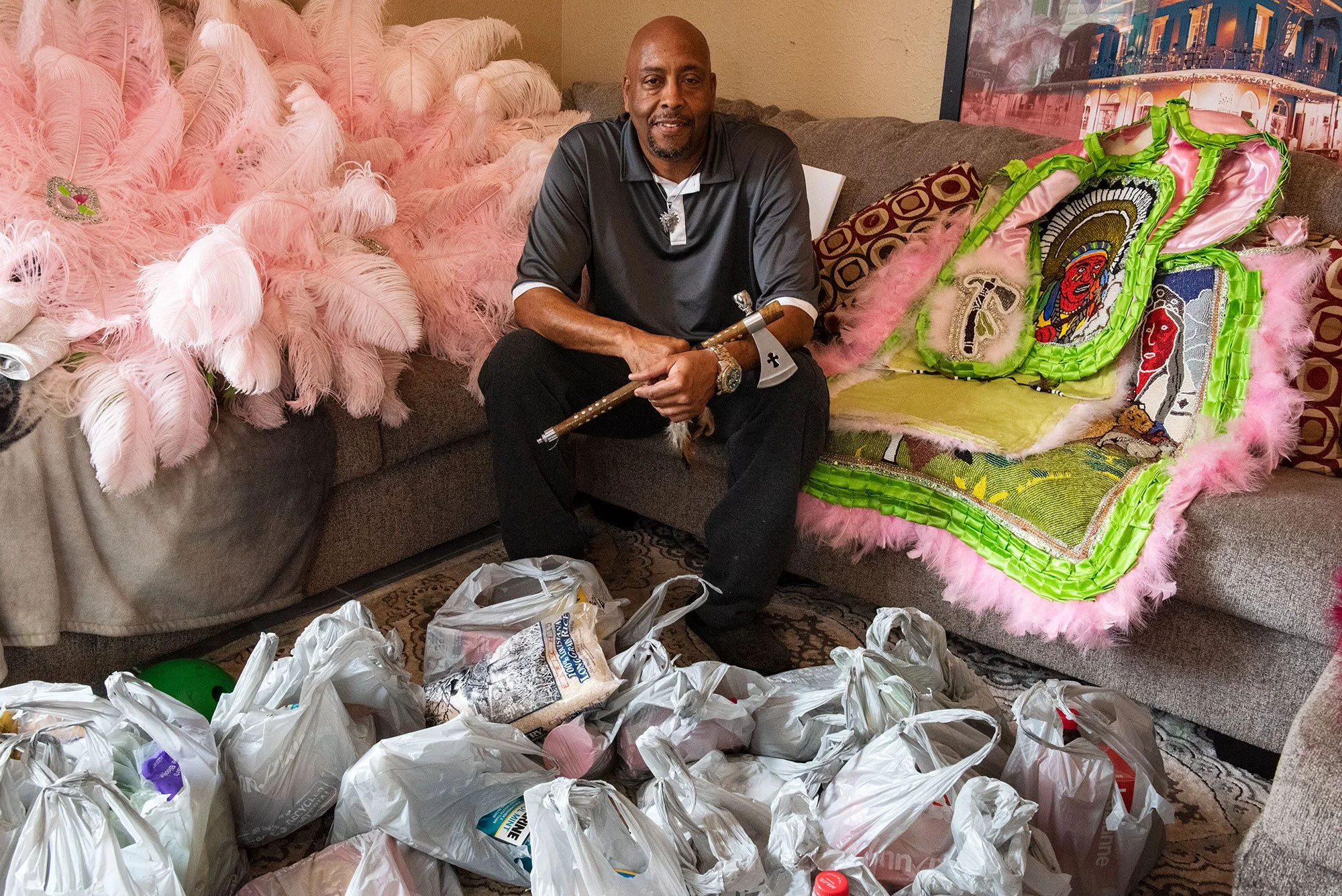 A man sitting on a beige couch holding a baton, surrounded by plastic bags on the floor, with colorful feathered and embroidered decorations on the couch beside him.