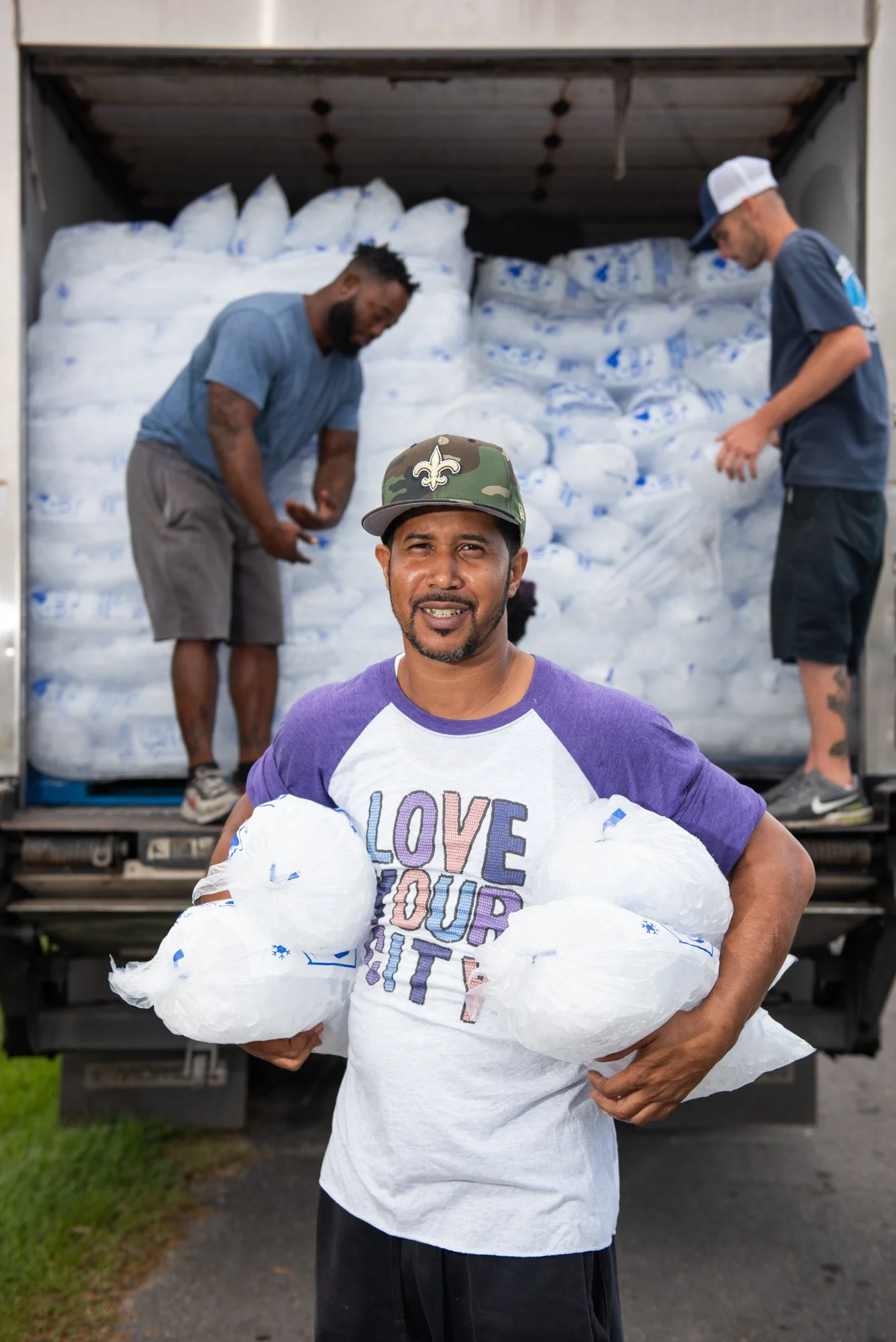 Three men unloading bags of supplies from a truck, one in the foreground holding large bags and wearing a shirt that says 'Love Your City'. The other two are in the truck, organizing or preparing more bags.