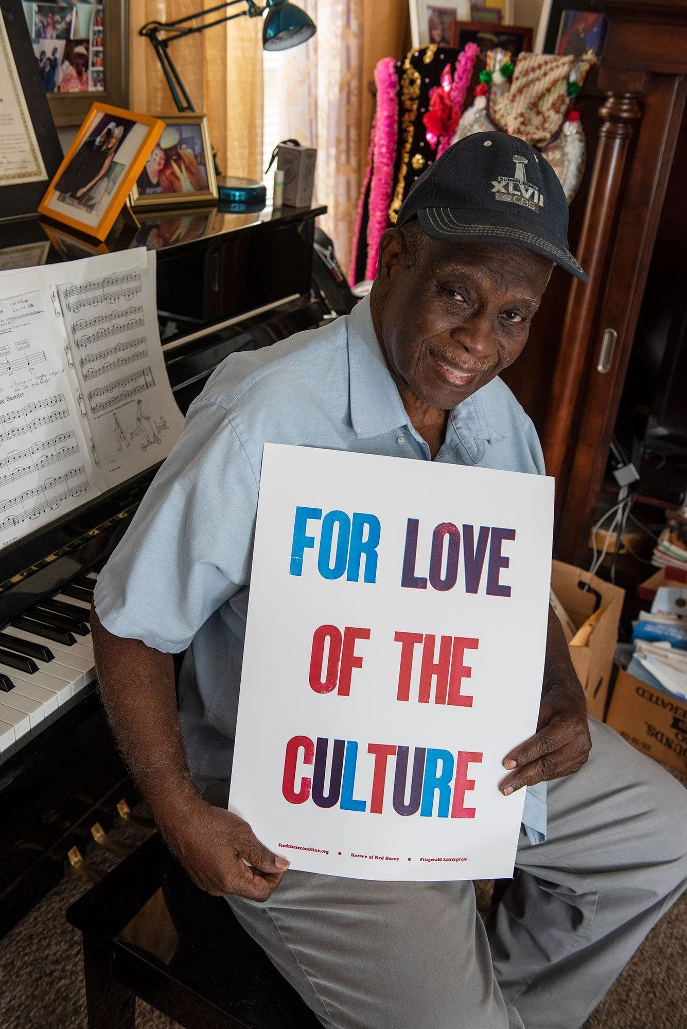 An older man sitting in front of a piano, holding a sign that reads 'For Love of the Culture' in colorful text, wearing a gray baseball cap and light blue shirt in a cozy room with photos and sheets of music around him.