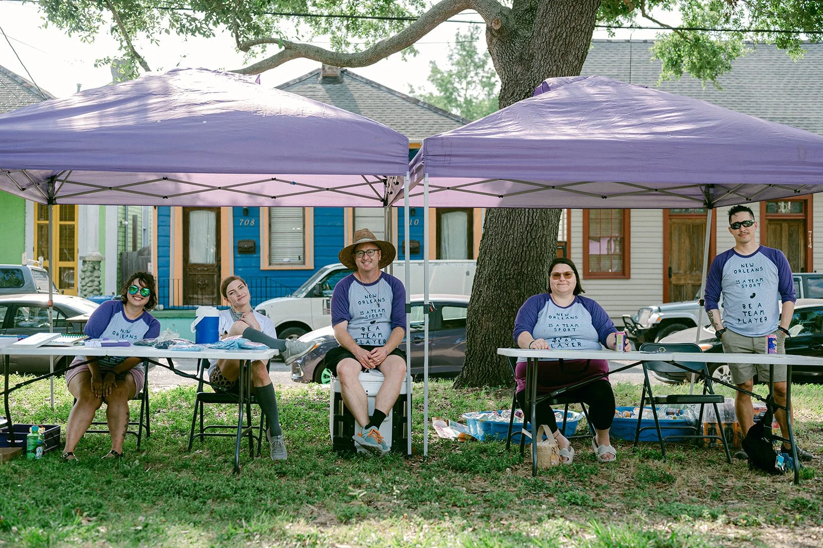 Five people sitting under two purple tents outdoors, with colorful houses in the background. All wearing matching shirts with the text "New Orleans is a team sport. Be a team player."