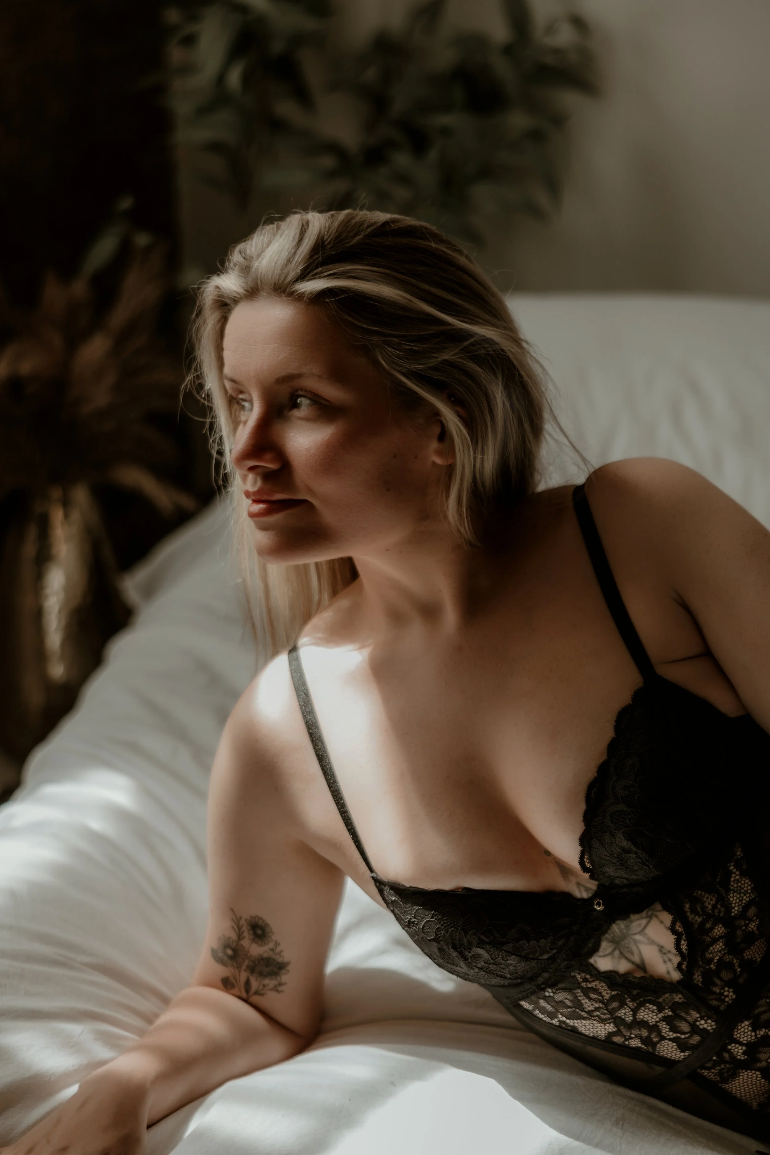 A woman reclining on a bed, wearing black lacy lingerie, with a tattoo on her left arm, looking to her right.