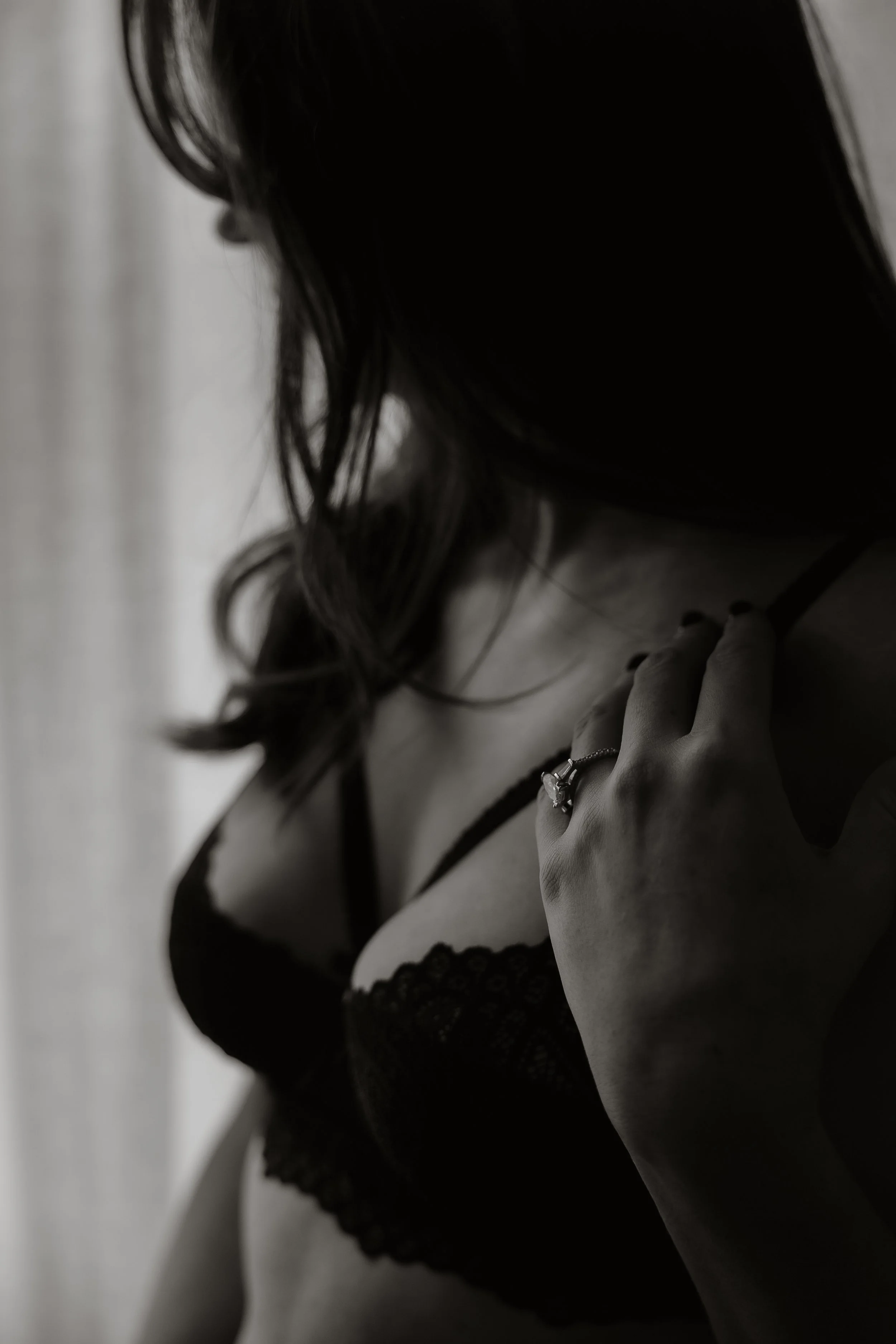 A woman in black lace lingerie with her hand on her collarbone, wearing a ring, in black and white.